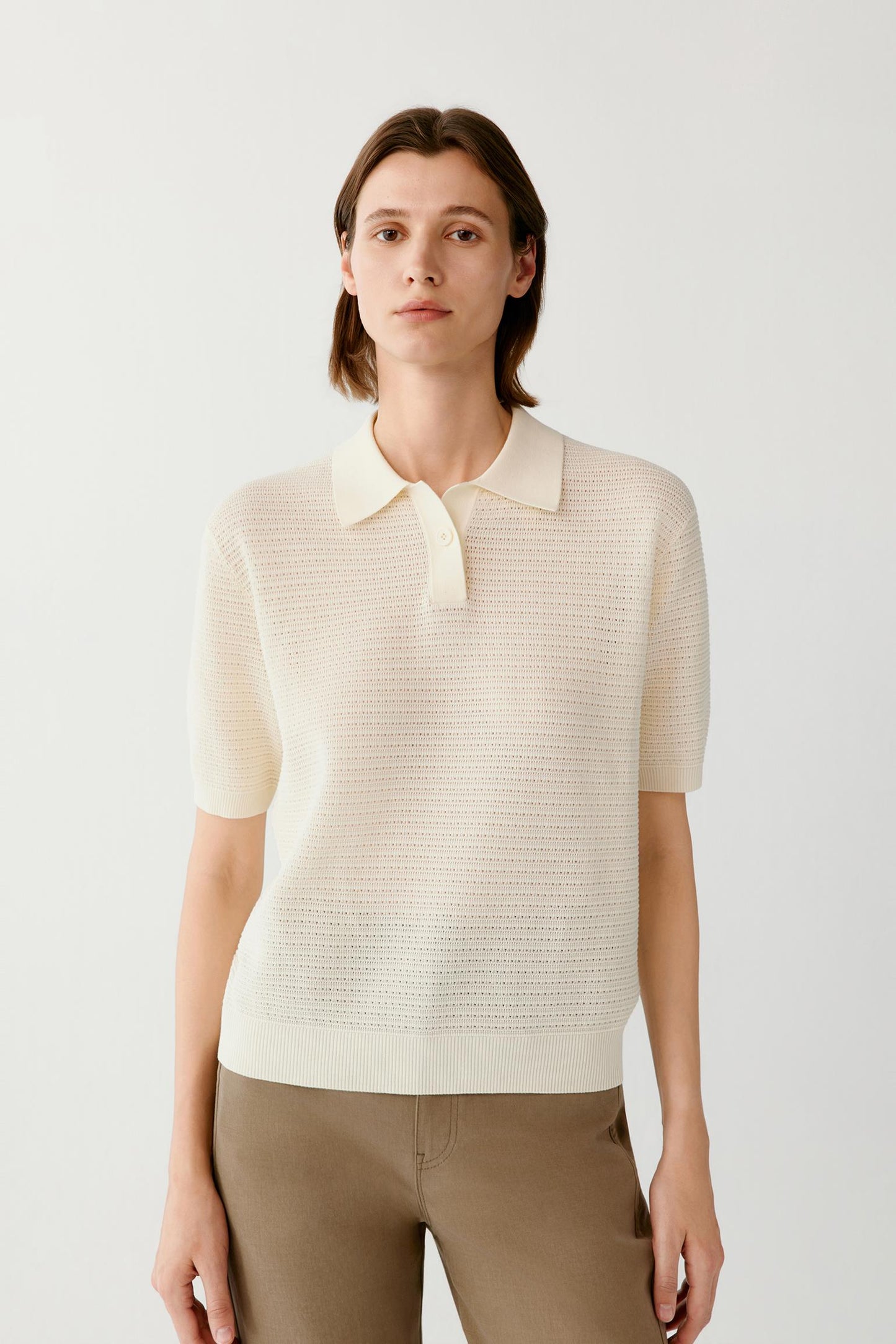 A woman modeling a short-sleeve knit polo shirt in cream, paired with brown trousers, against a plain light gray studio background.
