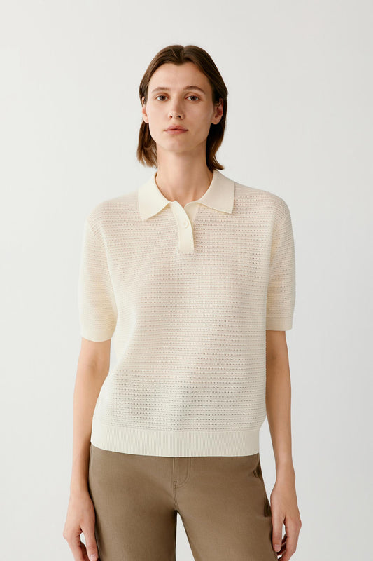A woman modeling a short-sleeve knit polo shirt in cream, paired with brown trousers, against a plain light gray studio background.