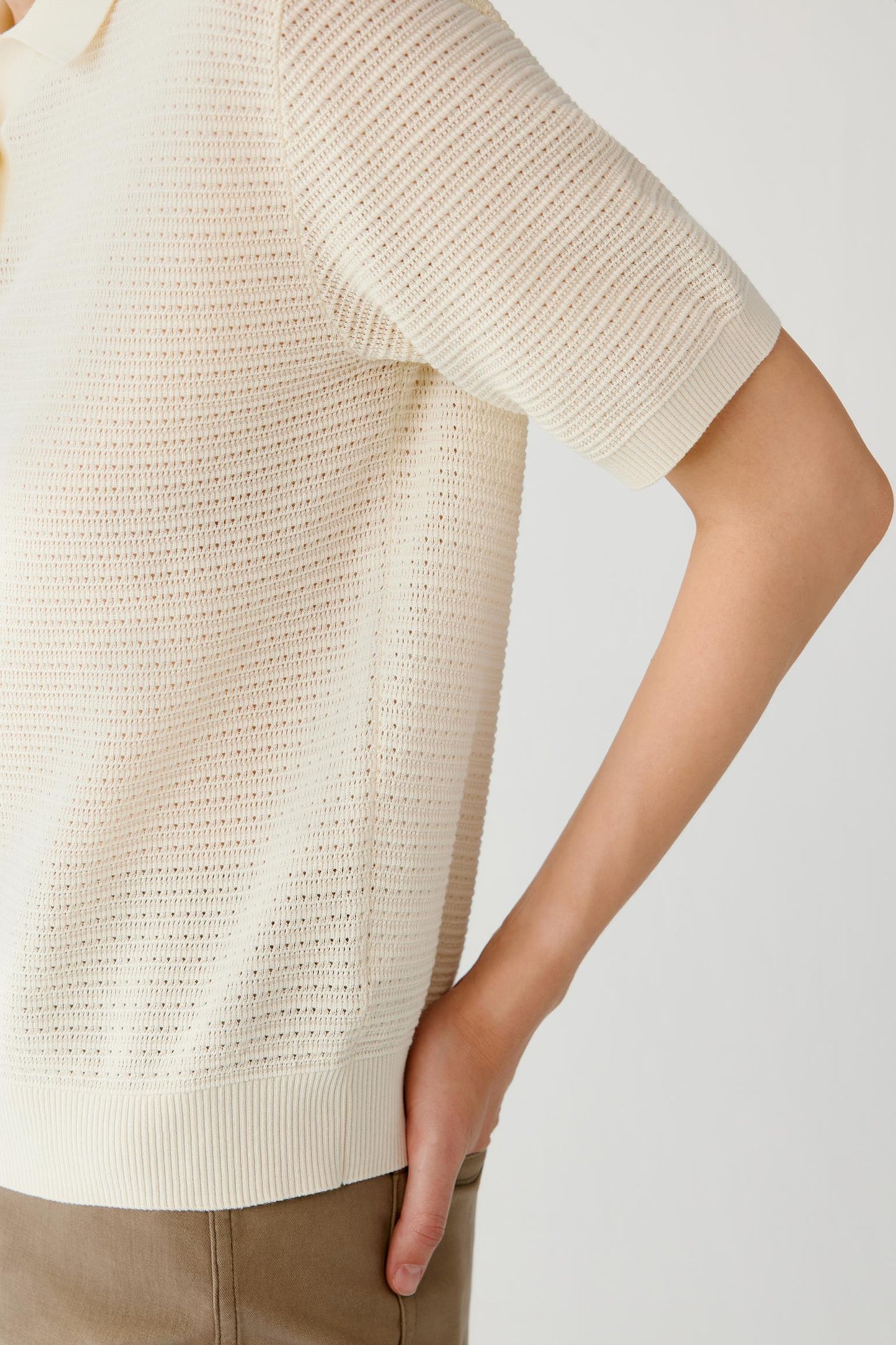 Close-up of a person wearing a short-sleeve cream knit sweater with a textured openwork pattern, paired with tan pants, against a light gray studio background.