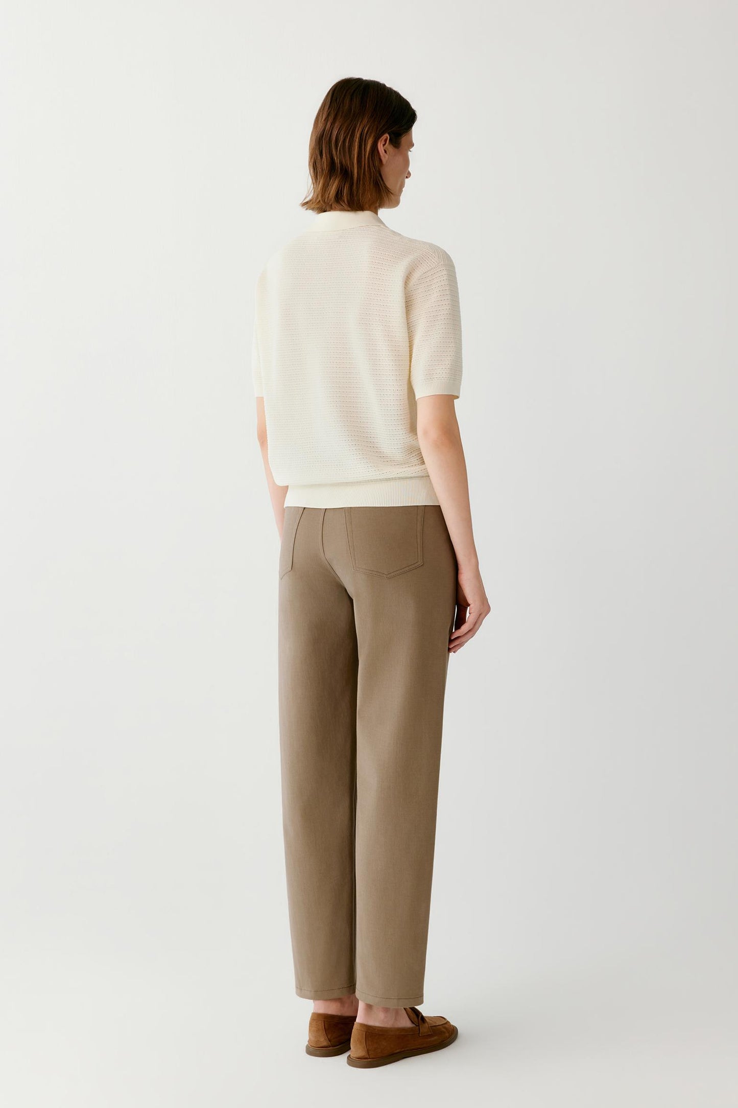 A model shown from the back wearing a cream short-sleeve knit top, beige wide-leg trousers, and brown suede loafers, set against a plain light gray studio background.