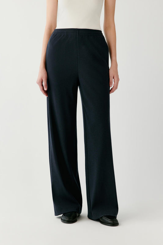 A model is shown from the waist down wearing a pair of dark navy wide-leg trousers, a cream sleeveless top, and black shoes against a light gray studio background.