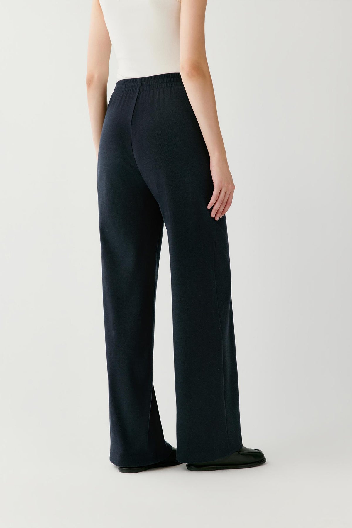 A model shown from the back wearing dark navy wide-leg trousers, a white sleeveless top, and black shoes against a light gray studio background.