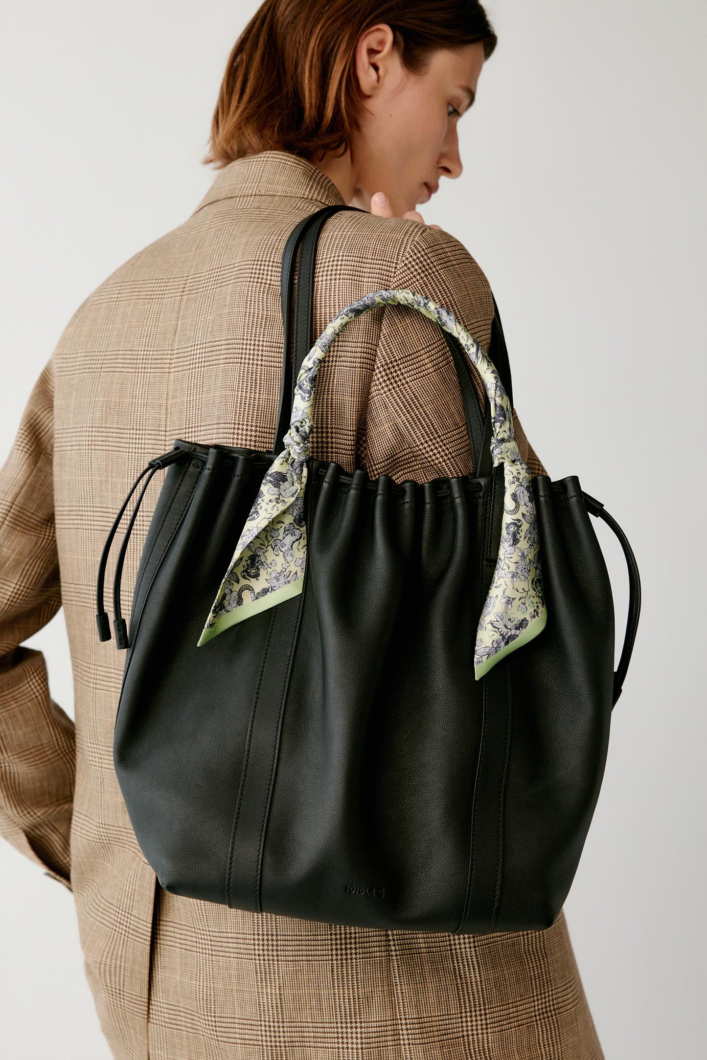 A woman wearing a beige plaid coat carries a black leather tote bag with a patterned scarf accent on the handles, photographed against a plain light gray background.