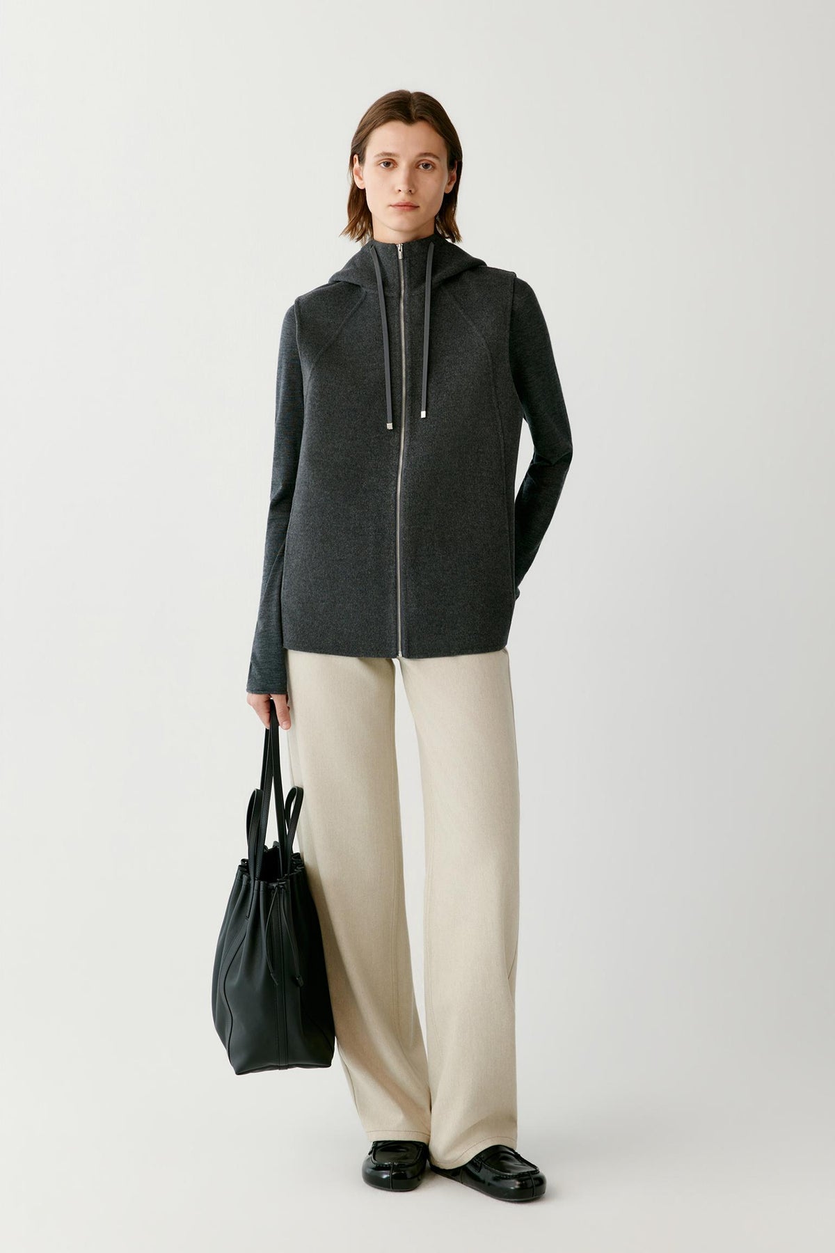 Front view of a woman wearing a dark grey melange sleeveless wool and cashmere coat, showing the hood, zip front and straight silhouette.