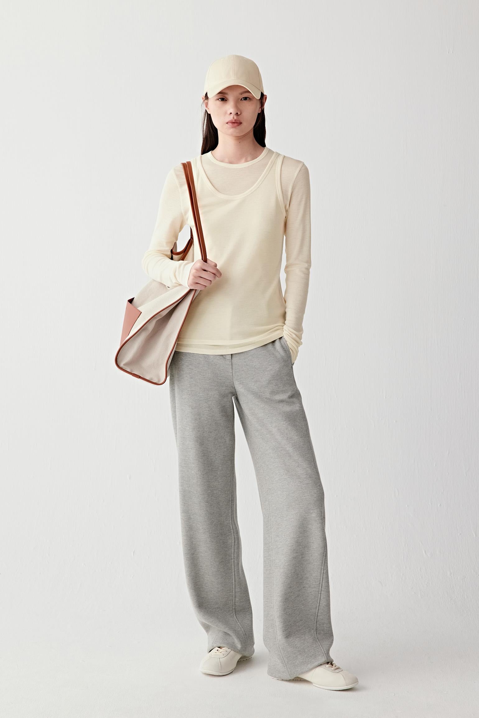 Person wearing a beige top and gray pants holding a brown bag on a white background