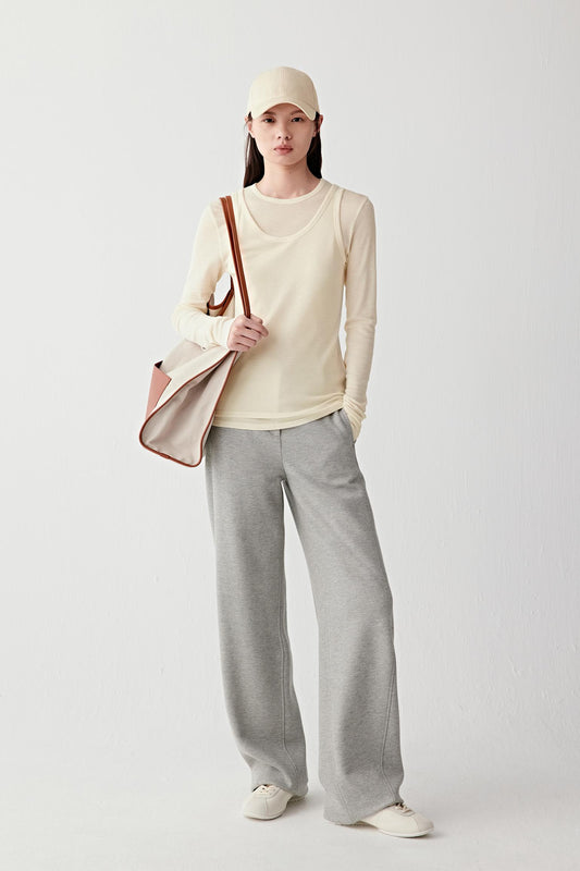 Person wearing a beige top and gray pants holding a brown bag on a white background