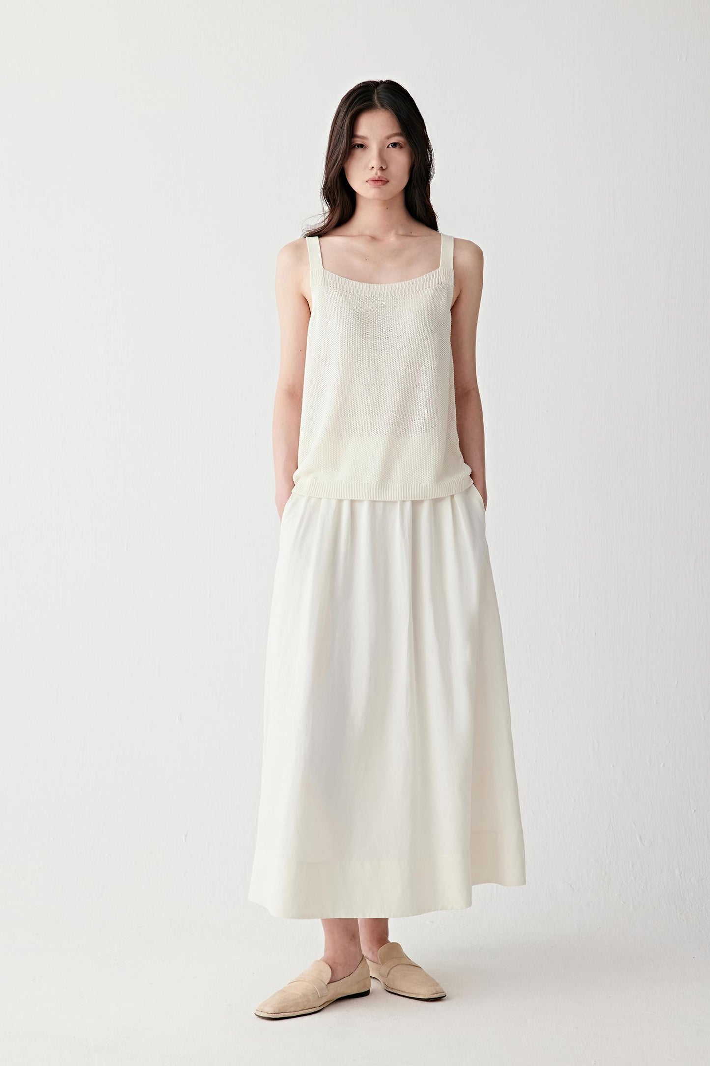 Full-length front view of a woman wearing a long off-white silk linen skirt with a gathered elastic waistband, showing soft volume, airy structure and fluid drape to the ankles, styled with a sleeveless top