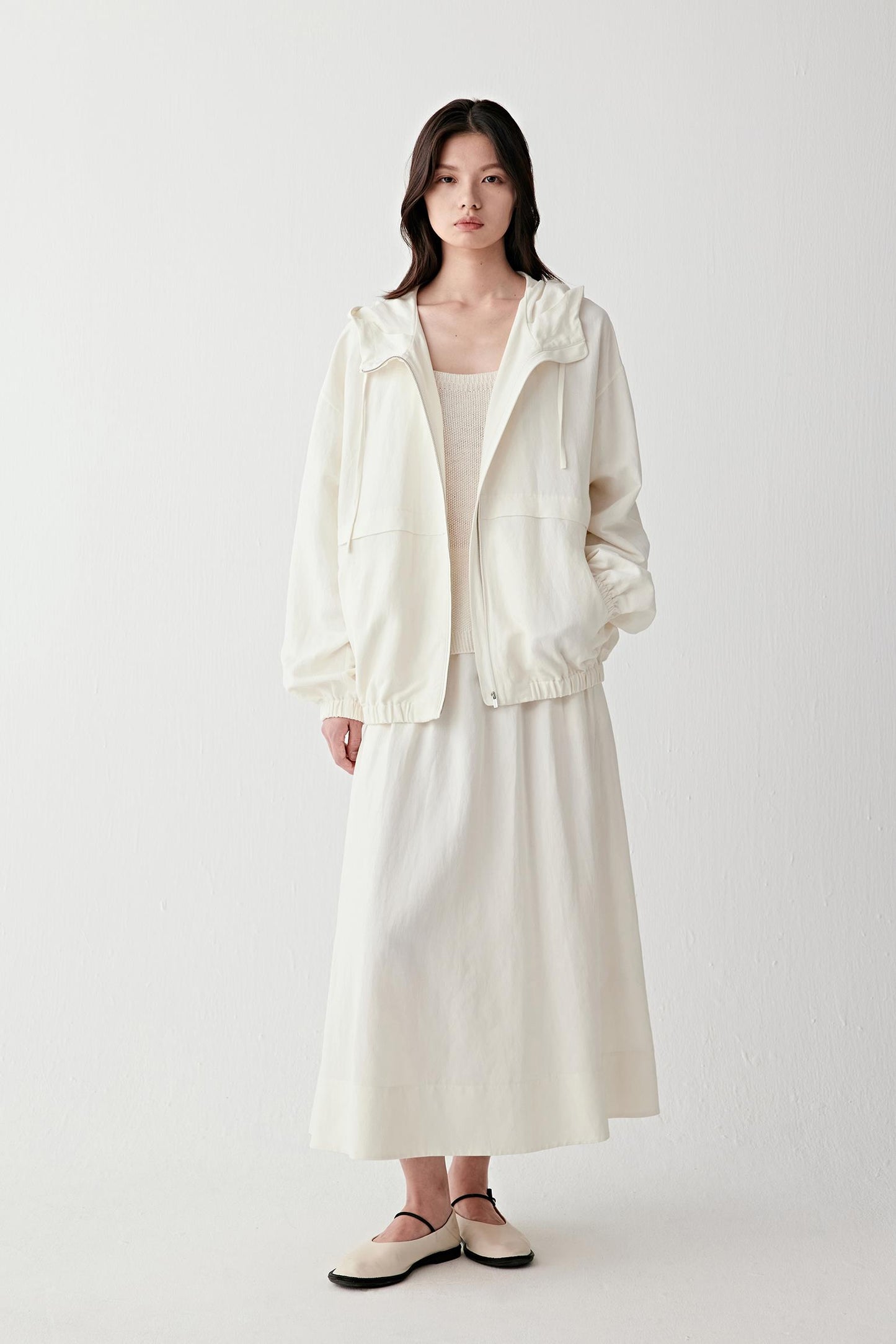 Full-length front view of a woman standing facing forward wearing an undyed white silk and linen jacket with a hood, paired with a matching long skirt, highlighting the relaxed silhouette, lightweight structure and softly textured surface of the fabric