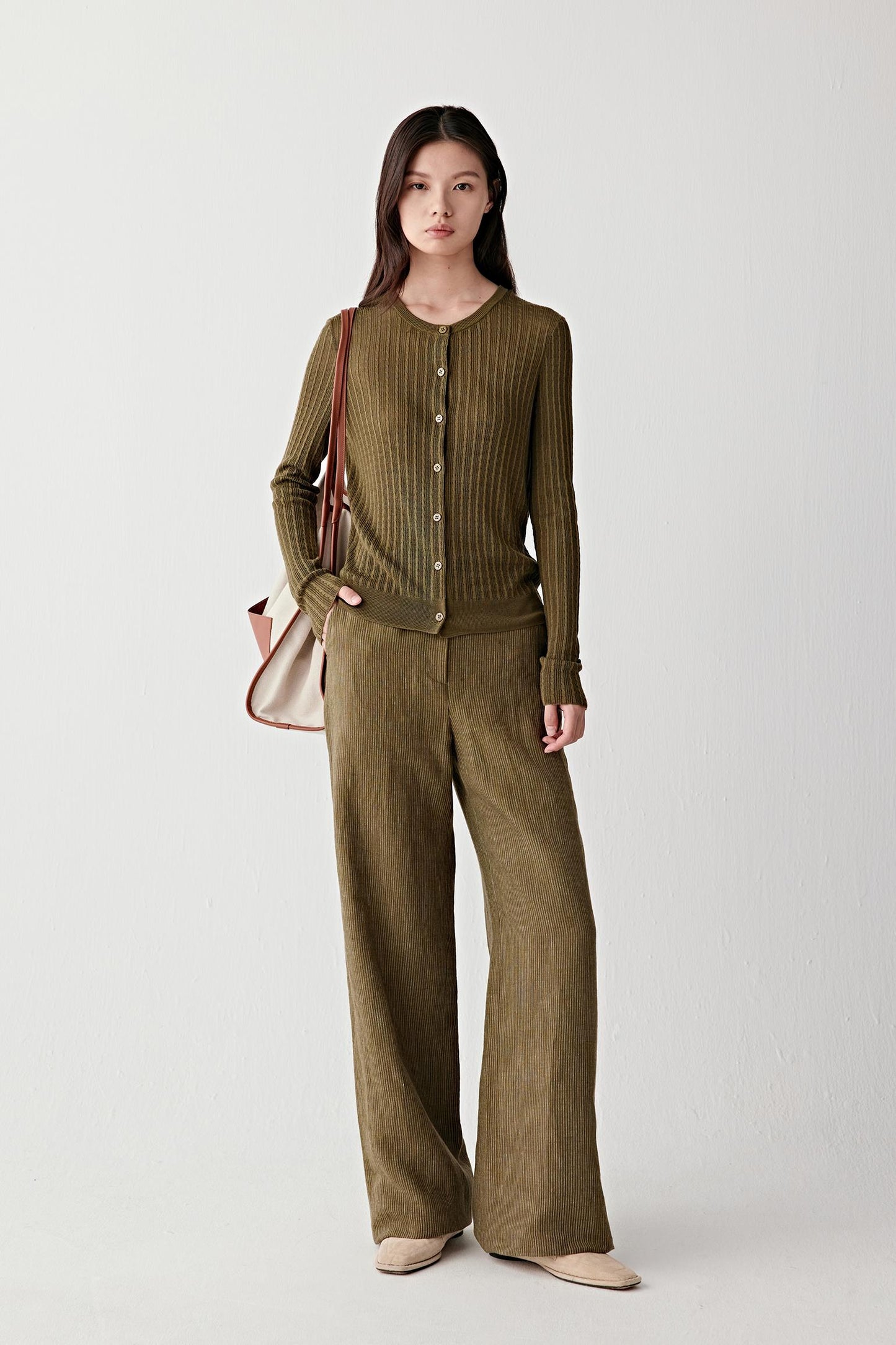 Full-length front view of a woman wearing olive green yarn-dyed linen trousers with a high waist and wide-leg cut, styled with a matching top, showing fluid drape, slightly textured surface and natural matte finish