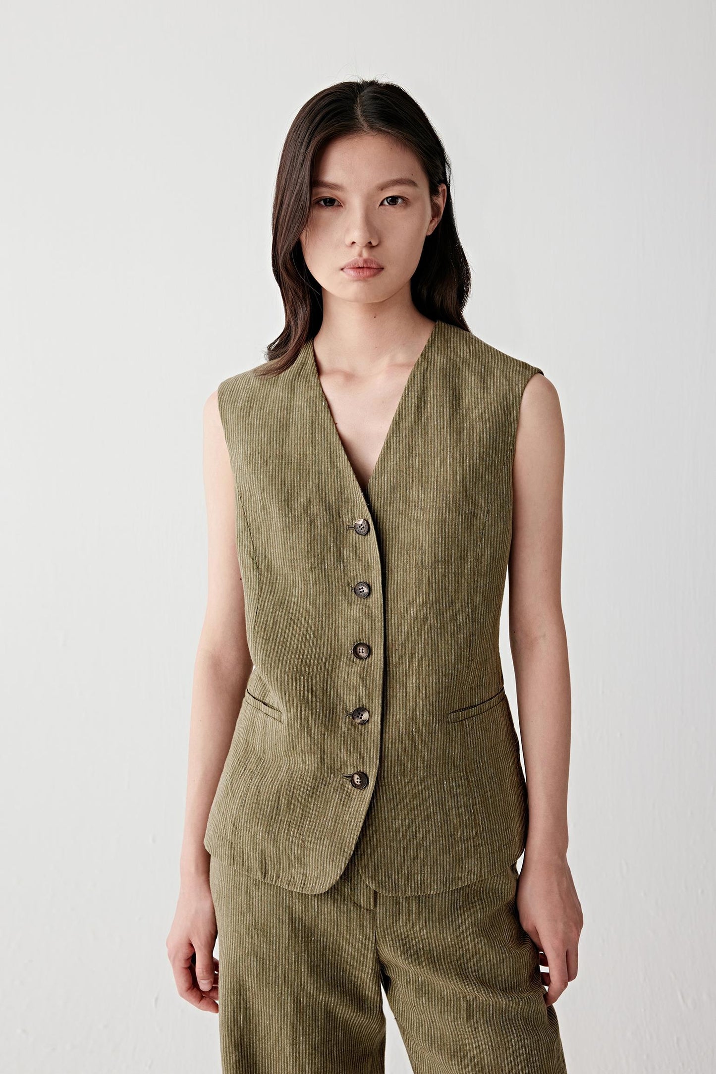 Front view portrait of a woman wearing a sleeveless khaki yarn-dyed linen vest with fine white stripes, highlighting the V-neckline, fitted silhouette and vertical stripe pattern that elongates the torso, with a lightly textured linen surface