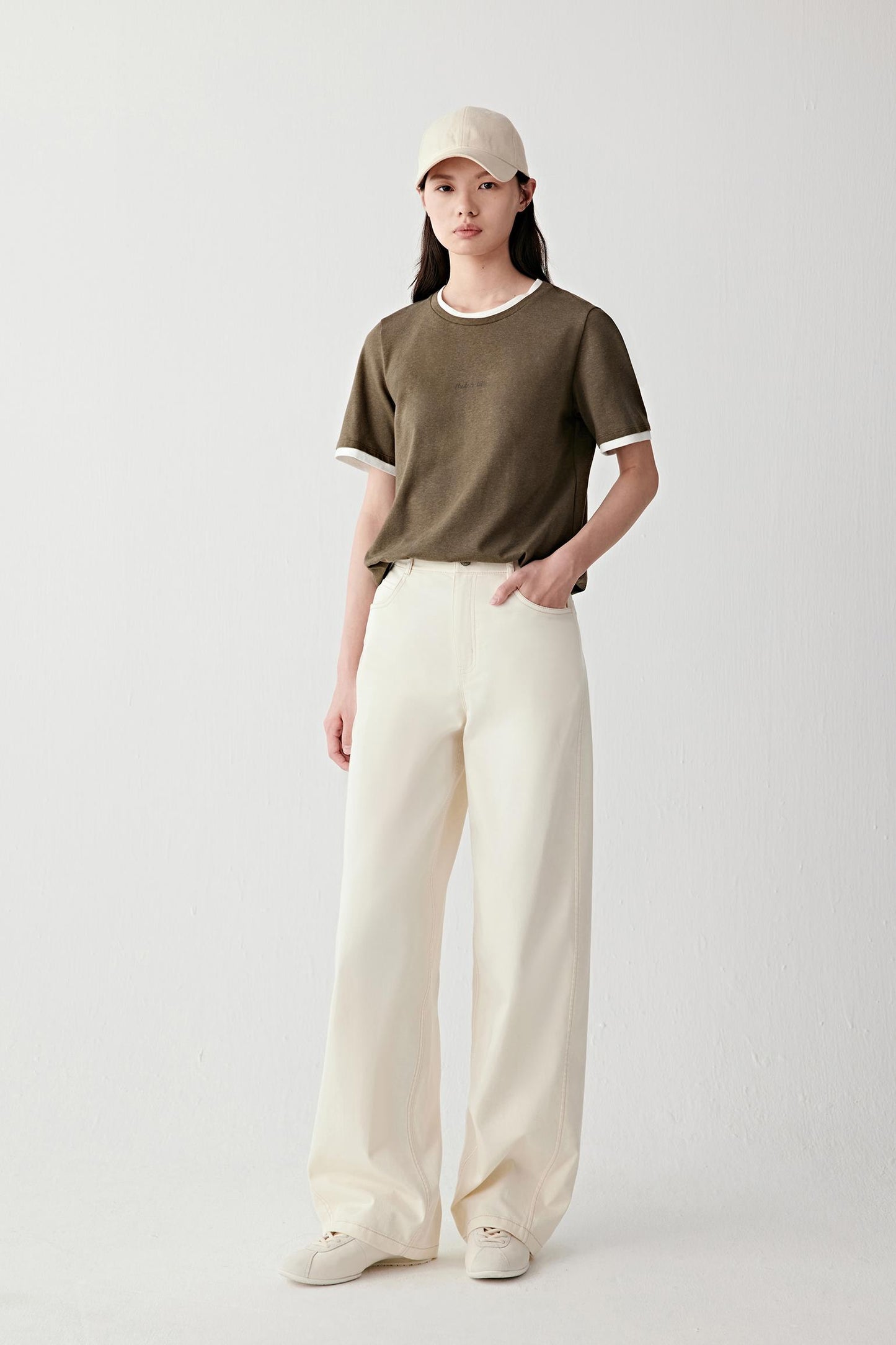 Full-length front view of a woman wearing ivory cotton stretch twill trousers with a high waist and wide-leg cut, styled with an olive T-shirt, showing structured yet fluid silhouette