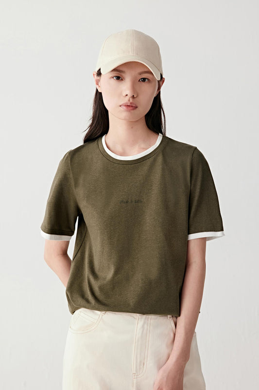 Three-quarter front view of a woman wearing an olive green cotton hemp jersey T-shirt with contrasting off-white neckline and sleeve edges, relaxed straight cut, soft drape and slightly textured surface, styled with light beige trousers and a cap