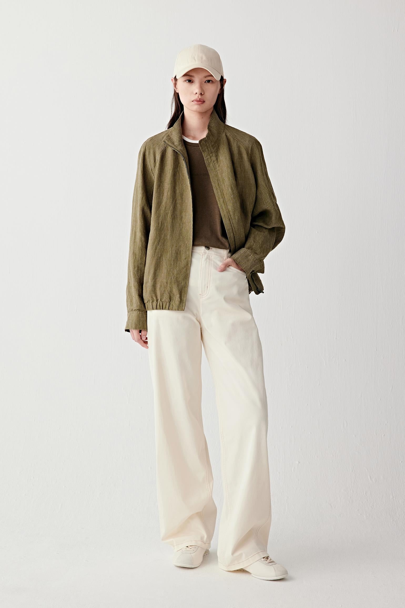 Full-length front view of a woman wearing an olive green yarn-dyed linen jacket with a relaxed, slightly oversized cut, paired with light trousers and a cap, showing lightweight structure and natural texture