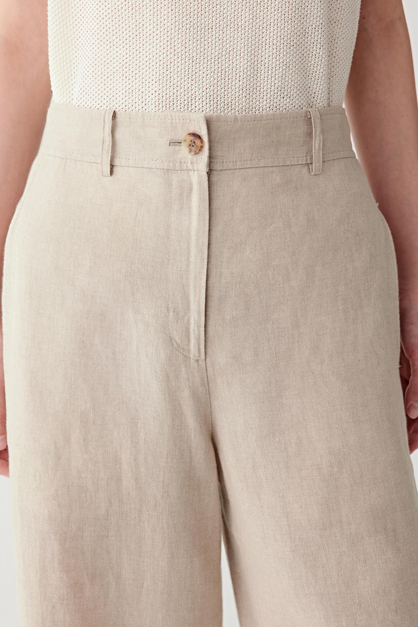 Close-up of waistband and pleats detailing the button fastening, front pleats and belt loops, highlighting the natural irregularities of the linen weave