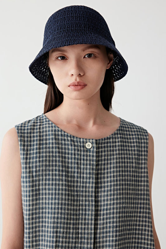 Front portrait view of a woman wearing a navy linen bucket hat, shown from chest up, highlighting the classic bucket shape, softly structured brim and textured openwork knit construction, paired with a sleeveless patterned top, with the hat sitting naturally on the head and casting a soft shadow over the face