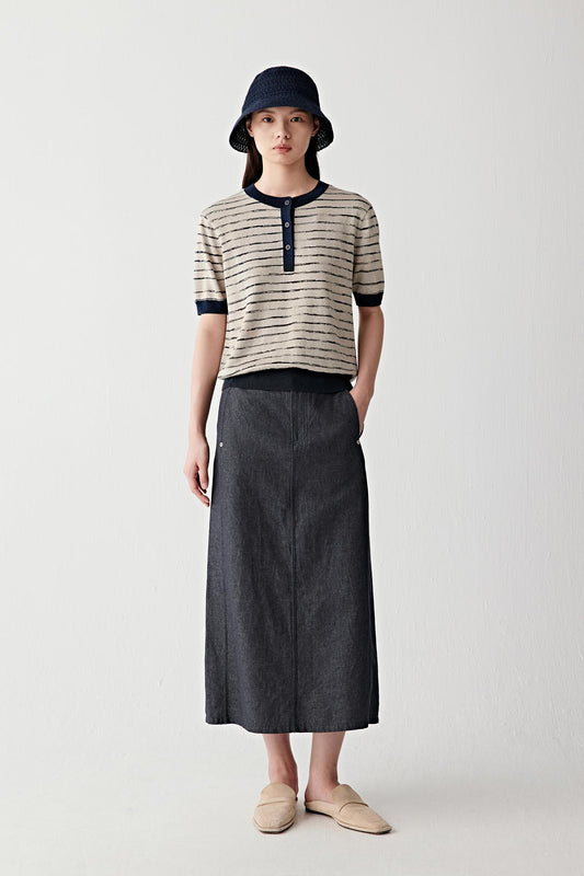 Full-length front view of a woman wearing a striped linen-silk pullover in beige and black horizontal stripes, paired with a dark skirt and bucket hat, showing relaxed fit and lightweight knit