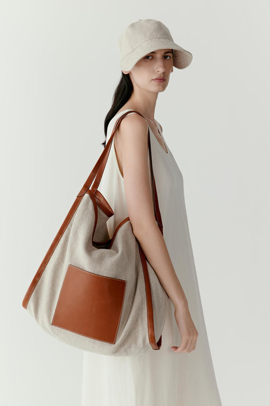 Front view of a model wearing a linen and cotton tote bag over the shoulder. The natural undyed fabric reveals subtle variations in tone, contrasted by a smooth camel leather pocket panel.