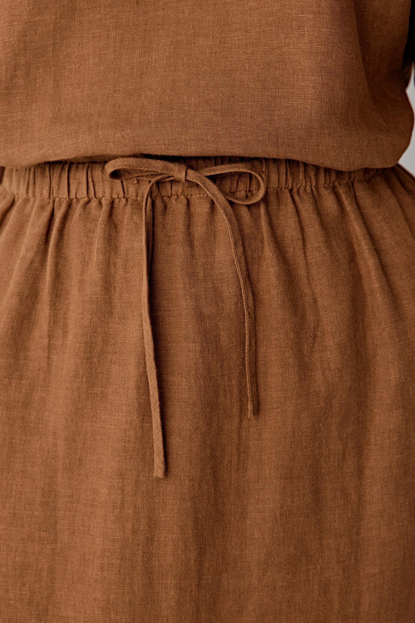 Close-up of the waistband and drawstring detailing the gathered construction, visible stitching and dry textured weave characteristic of plain-woven linen in a warm brown tone
