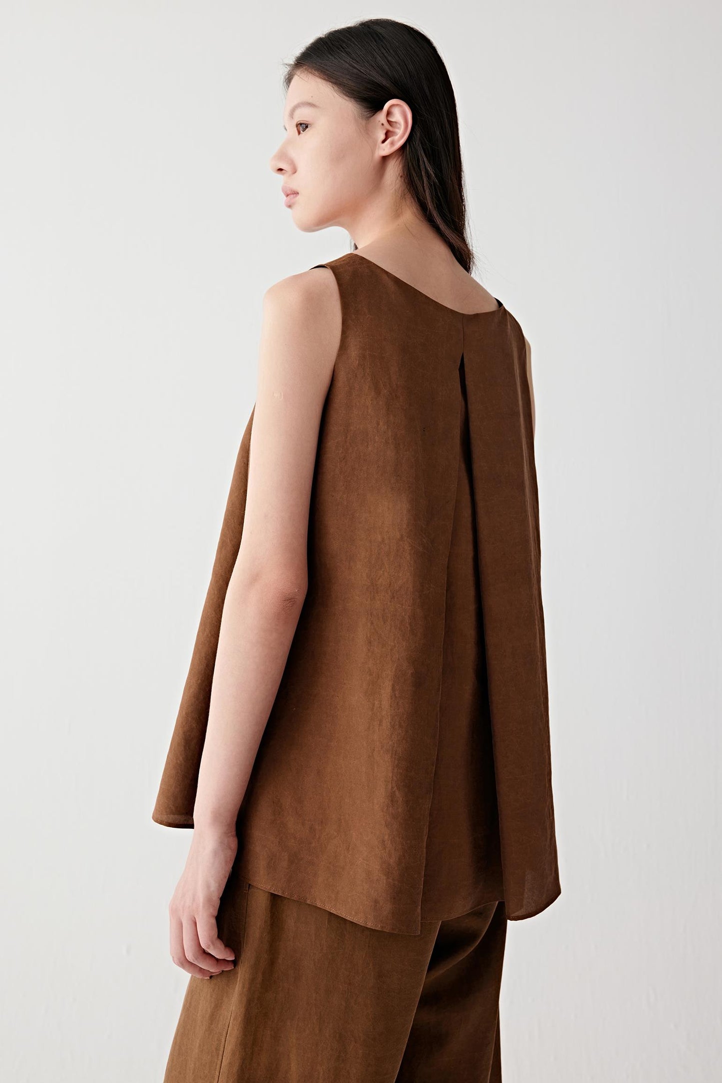 Three-quarter side or back view highlighting the clean sleeveless cut, back pleat construction and softly structured drape of the warm brown fabric