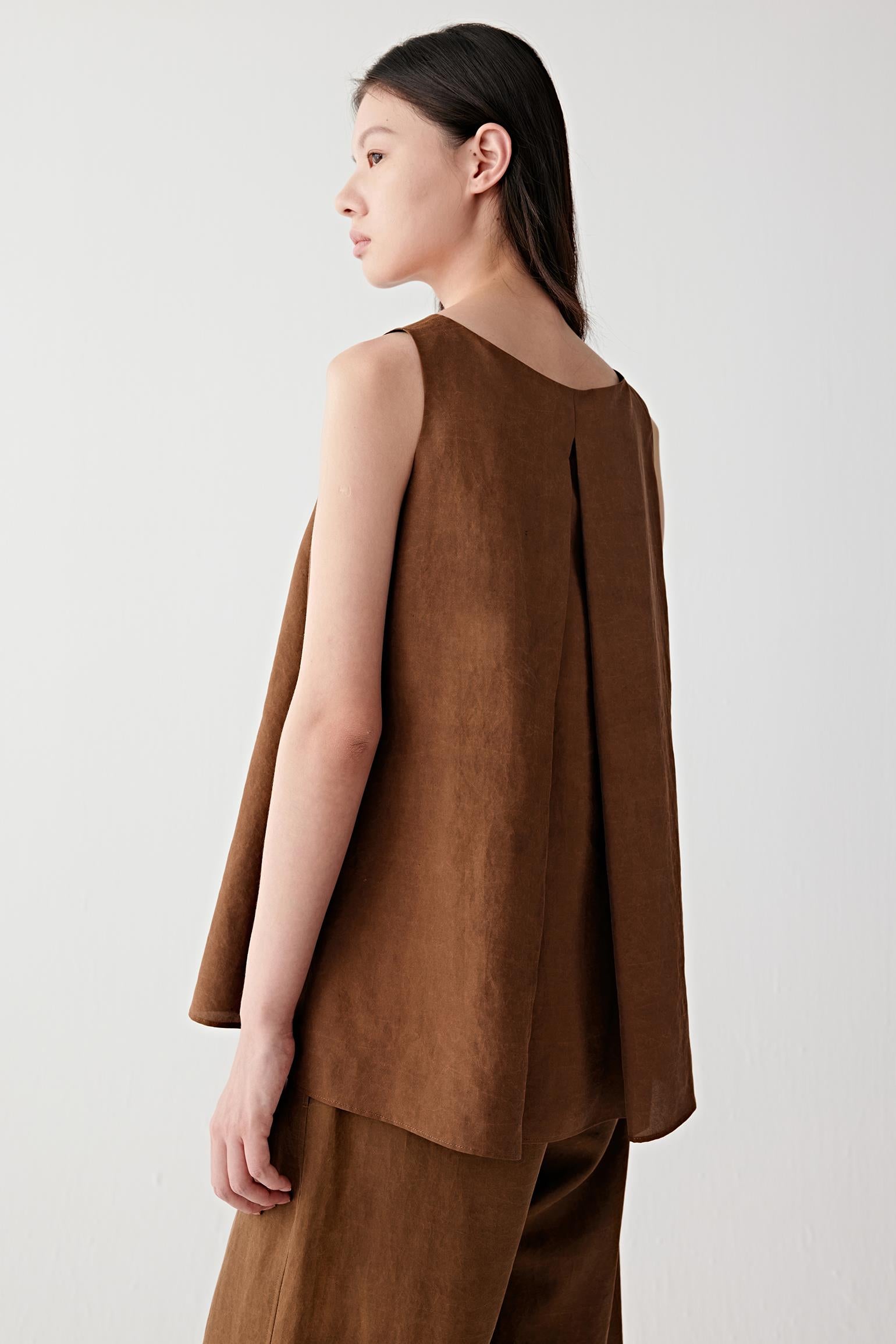 Three-quarter side or back view highlighting the clean sleeveless cut, back pleat construction and softly structured drape of the warm brown fabric
