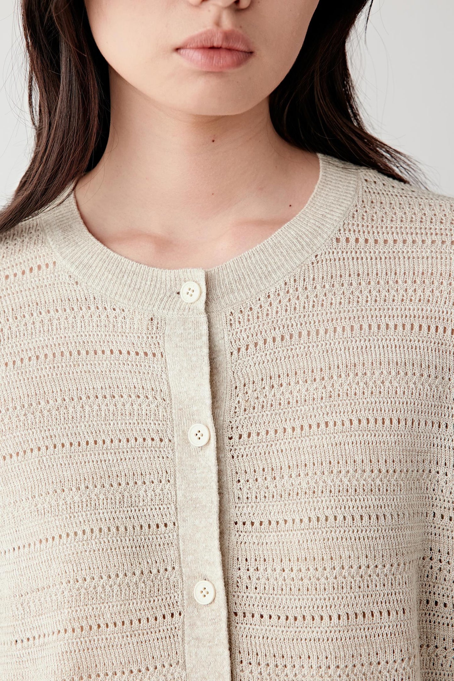 Close-up portrait focusing on the round neckline and button placket, detailing the alternating knit rows, perforated texture and softly textured beige melange yarn surface