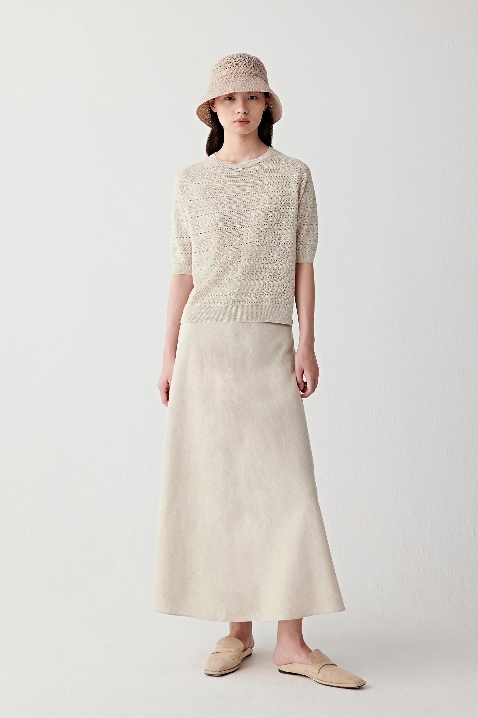Full-length front view of a woman wearing an undyed beige linen skirt paired with a matching sleeveless top and bucket hat, highlighting the lightweight woven structure, fluid drape and softly textured matte surface