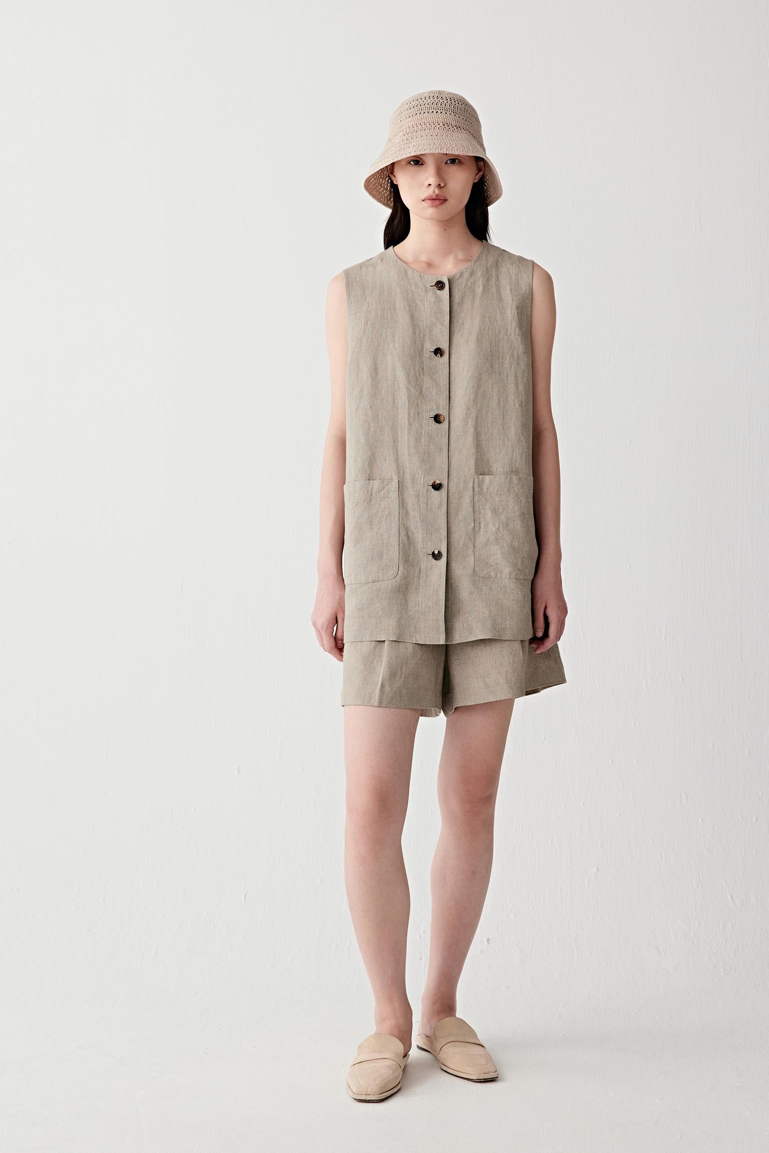 Full-length front view of a woman wearing a beige sleeveless linen jacket with a hooded silhouette, highlighting the dry textured surface, lightweight structure and softly straight fit