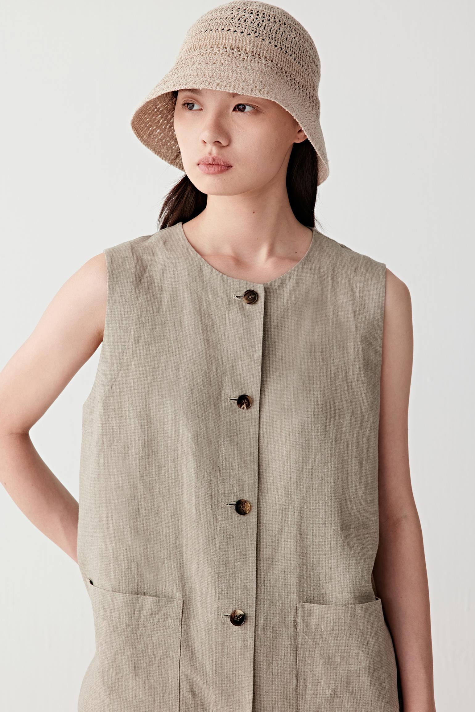Front portrait view of a woman wearing a beige melange linen and silk bucket hat, shown from chest up, highlighting the softly structured crown, gently sloping brim and openwork knit construction, with a lightly textured surface and subtle transparency, paired with a sleeveless linen top