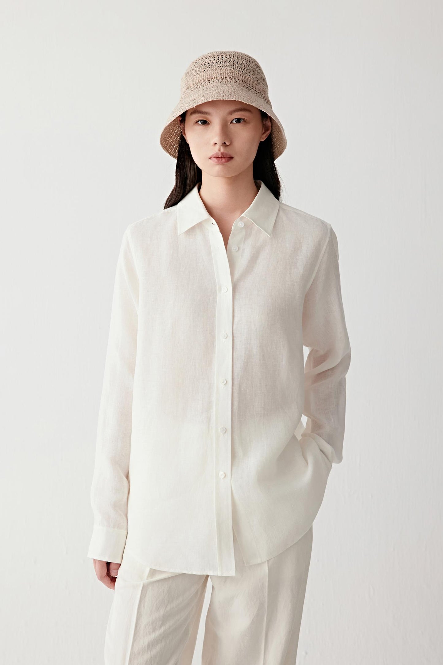Full-length front view of a woman wearing a light cream linen shirt with a relaxed silhouette, highlighting the lightweight woven structure, fluid drape and softly irregular linen surface