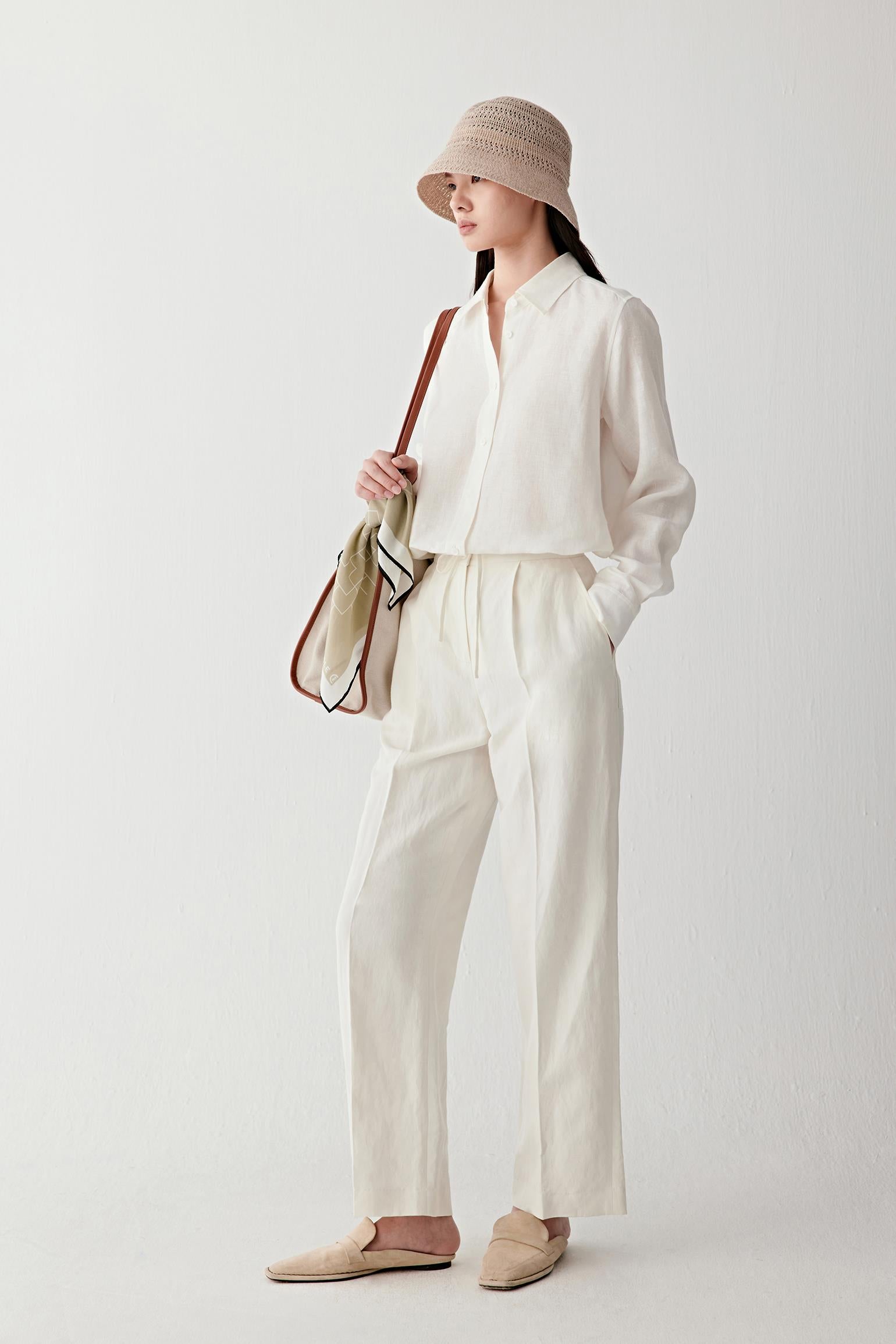 Full-length front view of a woman wearing undyed silk and linen trousers with a straight-leg silhouette, paired with a lightweight matching top and hat, highlighting the fluid drape, natural texture and soft matte surface of the fabric blend