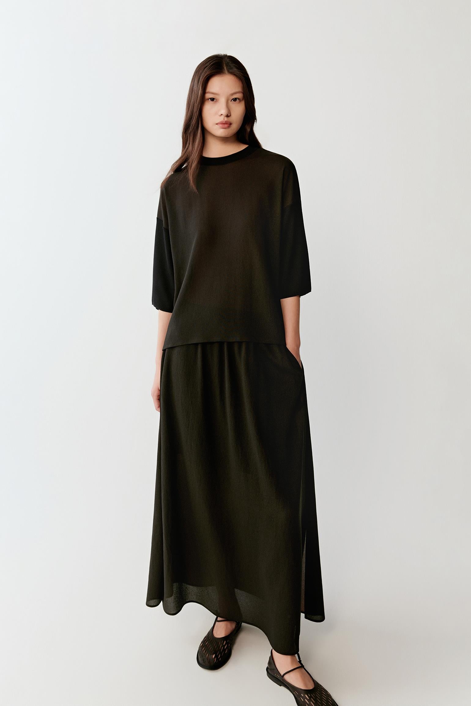 Full-length front view showing the pullover paired with a matching black skirt, emphasizing the elongated silhouette, fluid movement and matte silk surface with a soft hand