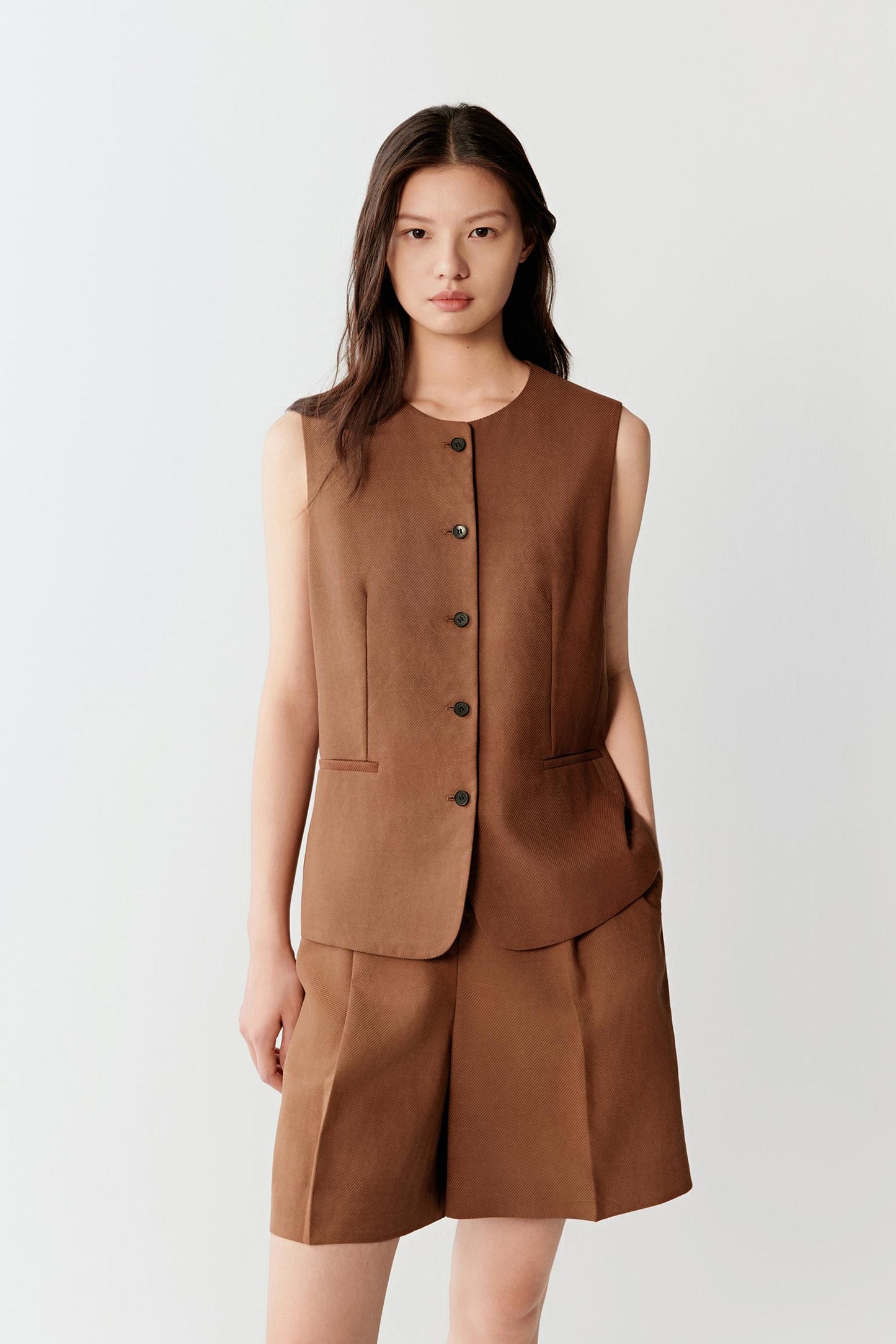 Front view of a woman wearing a warm brown Gambiered Canton Gauze vest with a sleeveless tailored silhouette, highlighting the compact woven structure, matte textured surface and softly structured drape developed through the traditional dyeing process