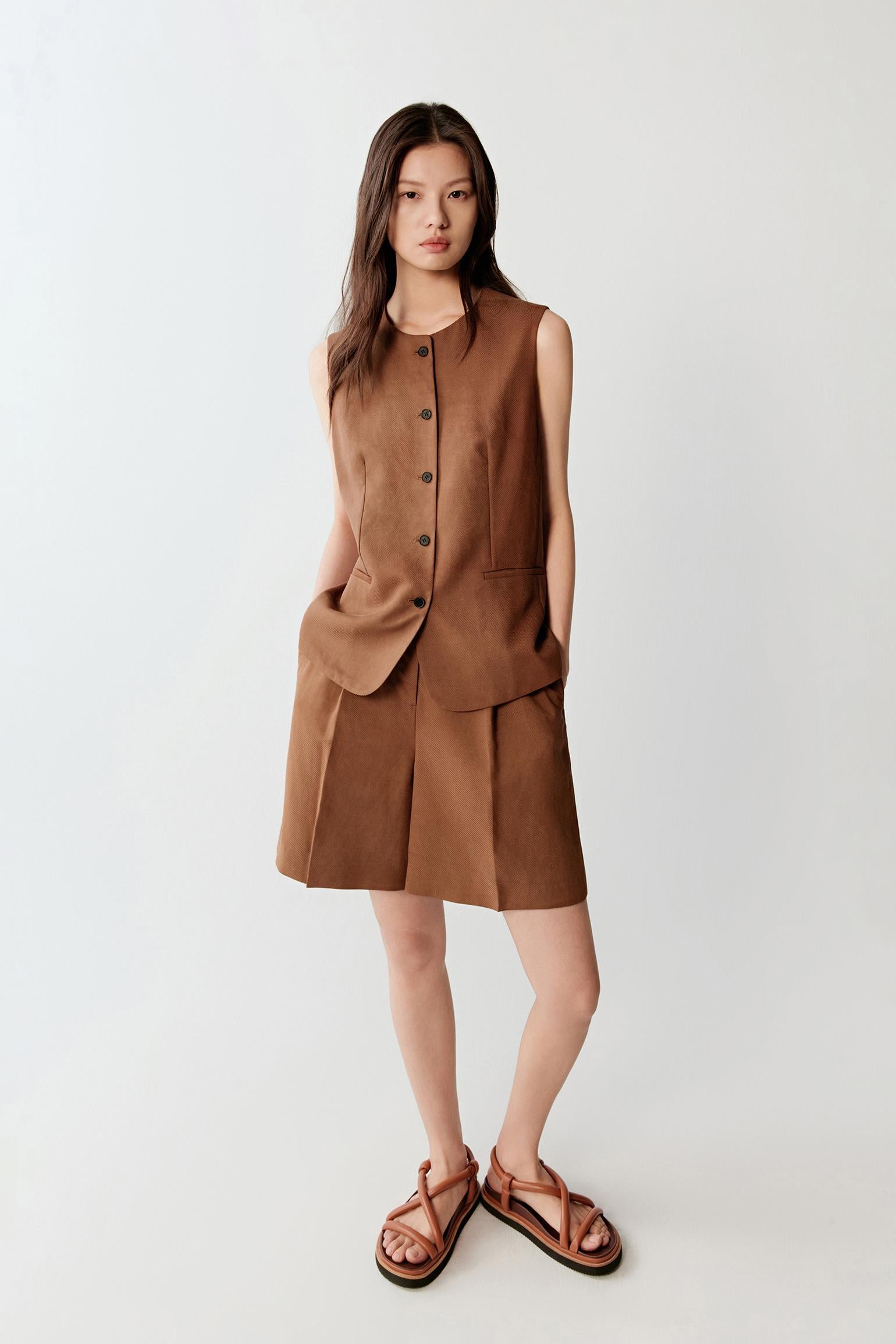 Full-length front view of a woman wearing warm brown Gambiered Canton Gauze shorts paired with a sleeveless black top, highlighting the structured silhouette, lightweight woven texture and matte surface with tonal depth