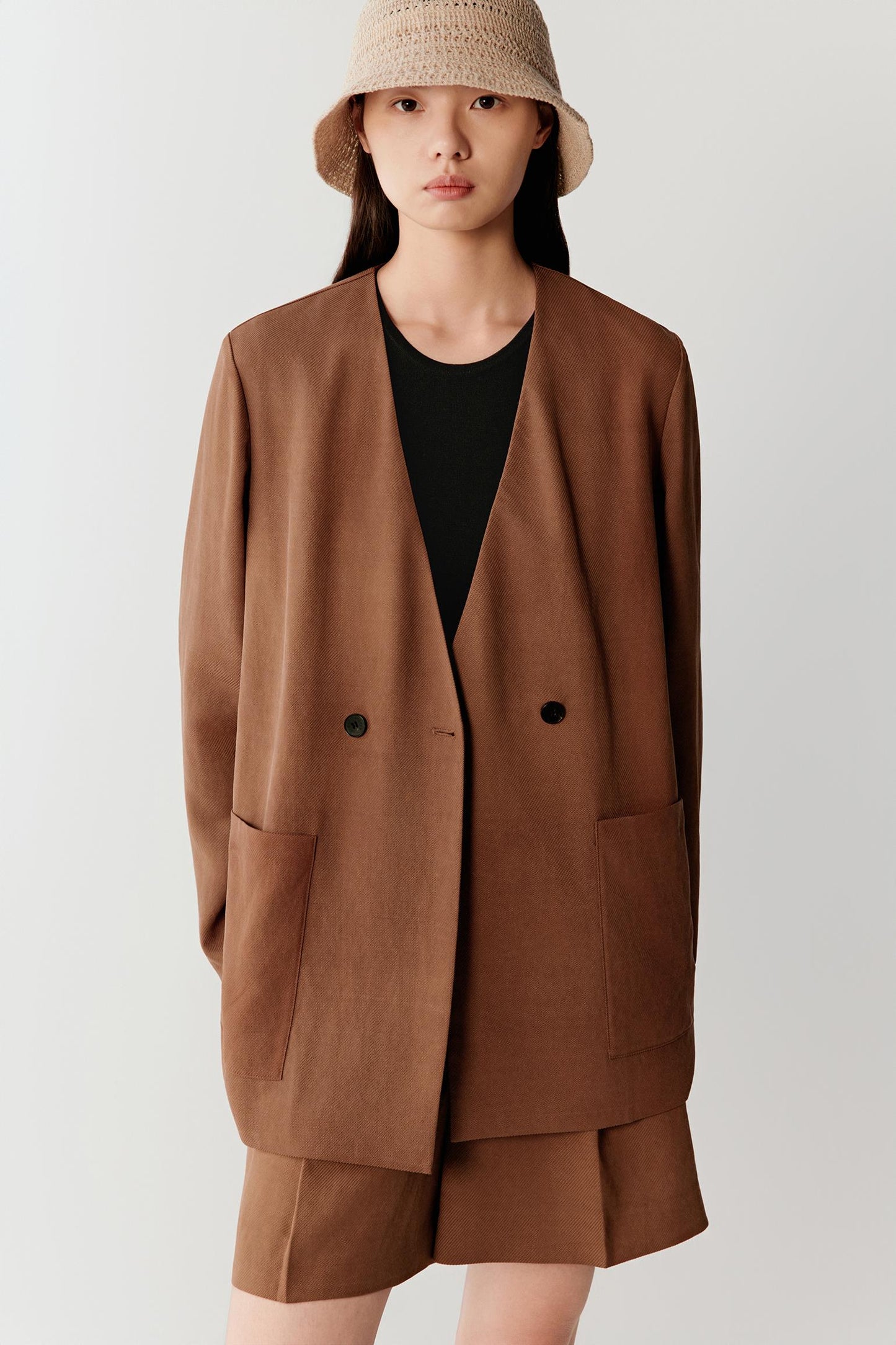 Front view of a woman wearing a warm brown Gambiered Canton Gauze jacket with an oversized silhouette and double-breasted fastening, highlighting the smooth matte surface, fluid drape and softly structured shape