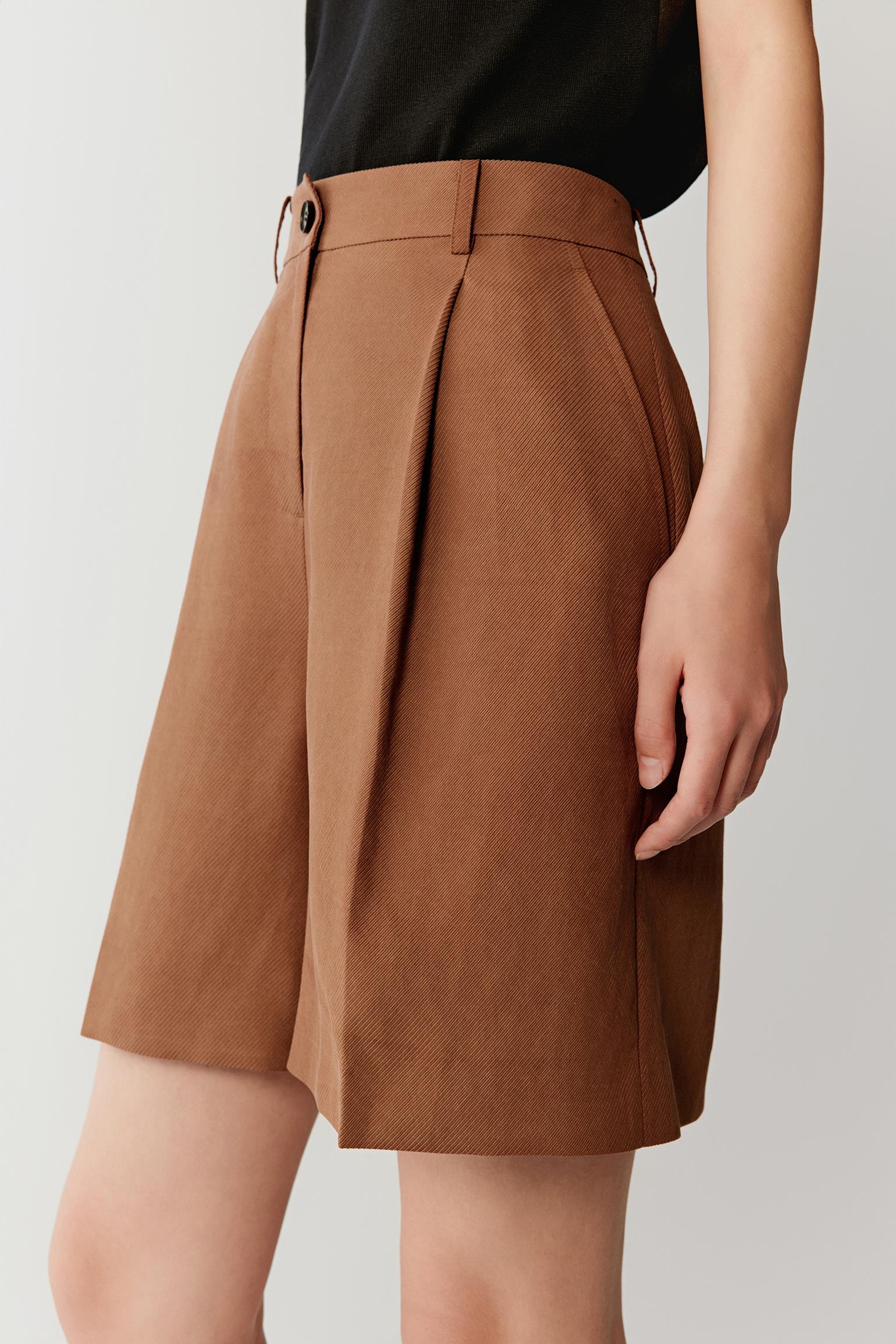 Close-up of waistband and pleats detailing the button fastening, belt loops, front pleat construction and subtle tonal variations in the warm brown fabric surface