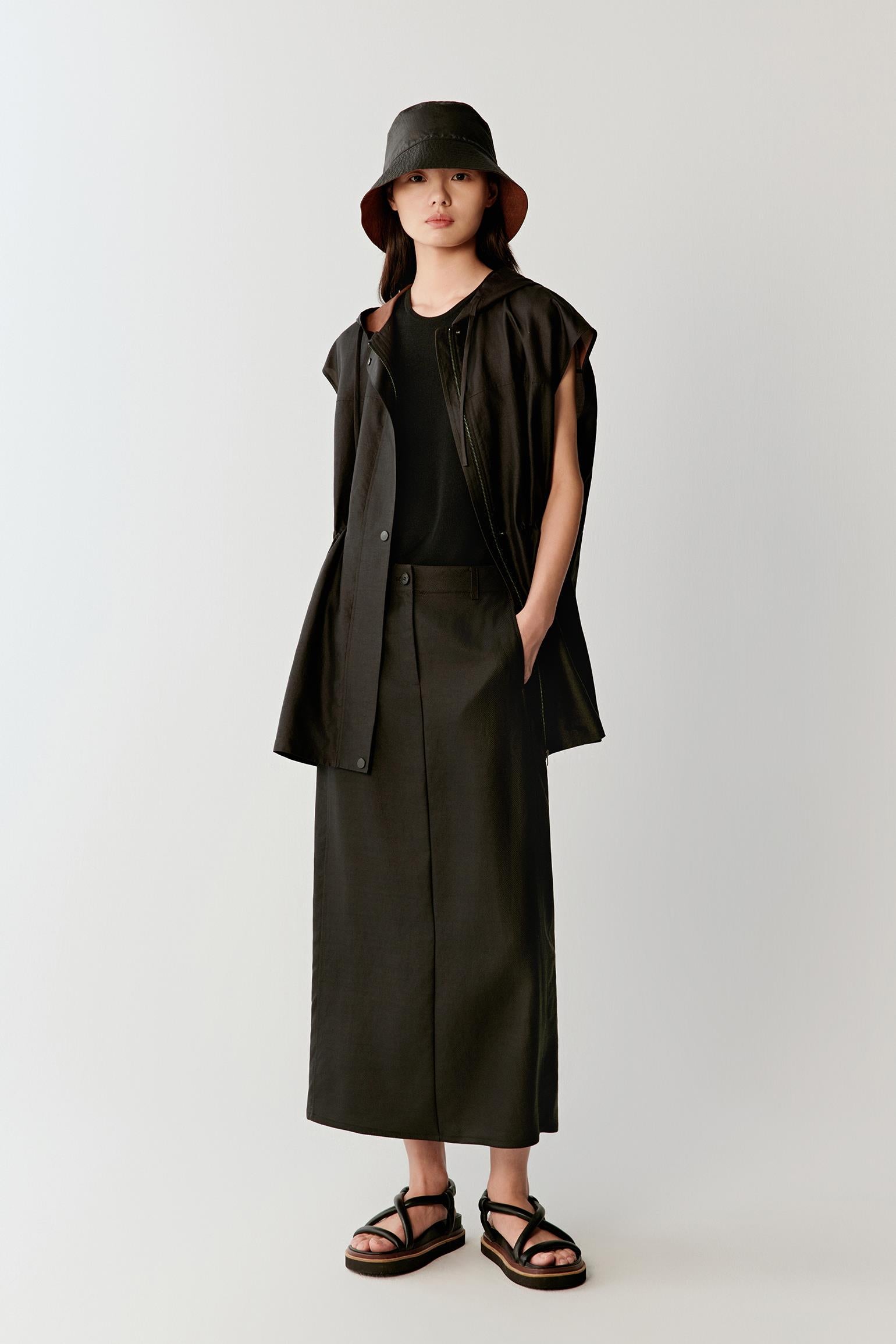 Full-length front view of a woman wearing a deep black sleeveless Gambiered Canton Gauze jacket with a fluid elongated silhouette, highlighting the hooded construction, lightweight drape and softly structured matte surface