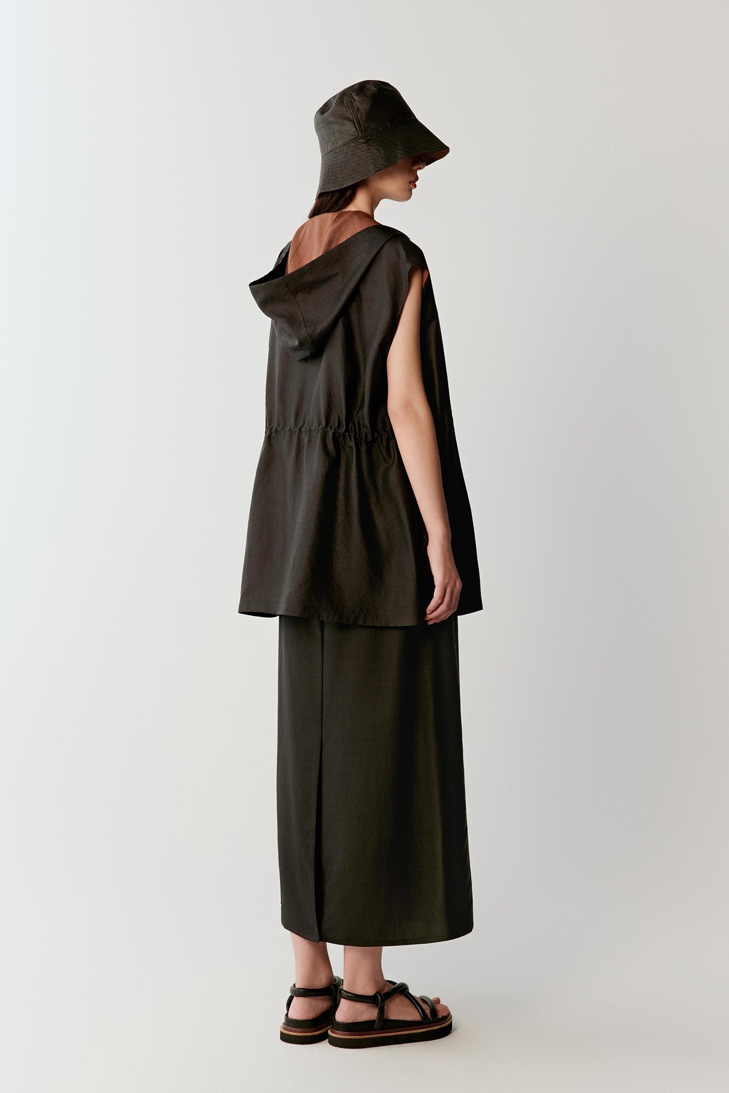 Full-length side or back view highlighting the elongated line, sleeveless construction and softly flowing drape of the deep black fabric as it moves around the body