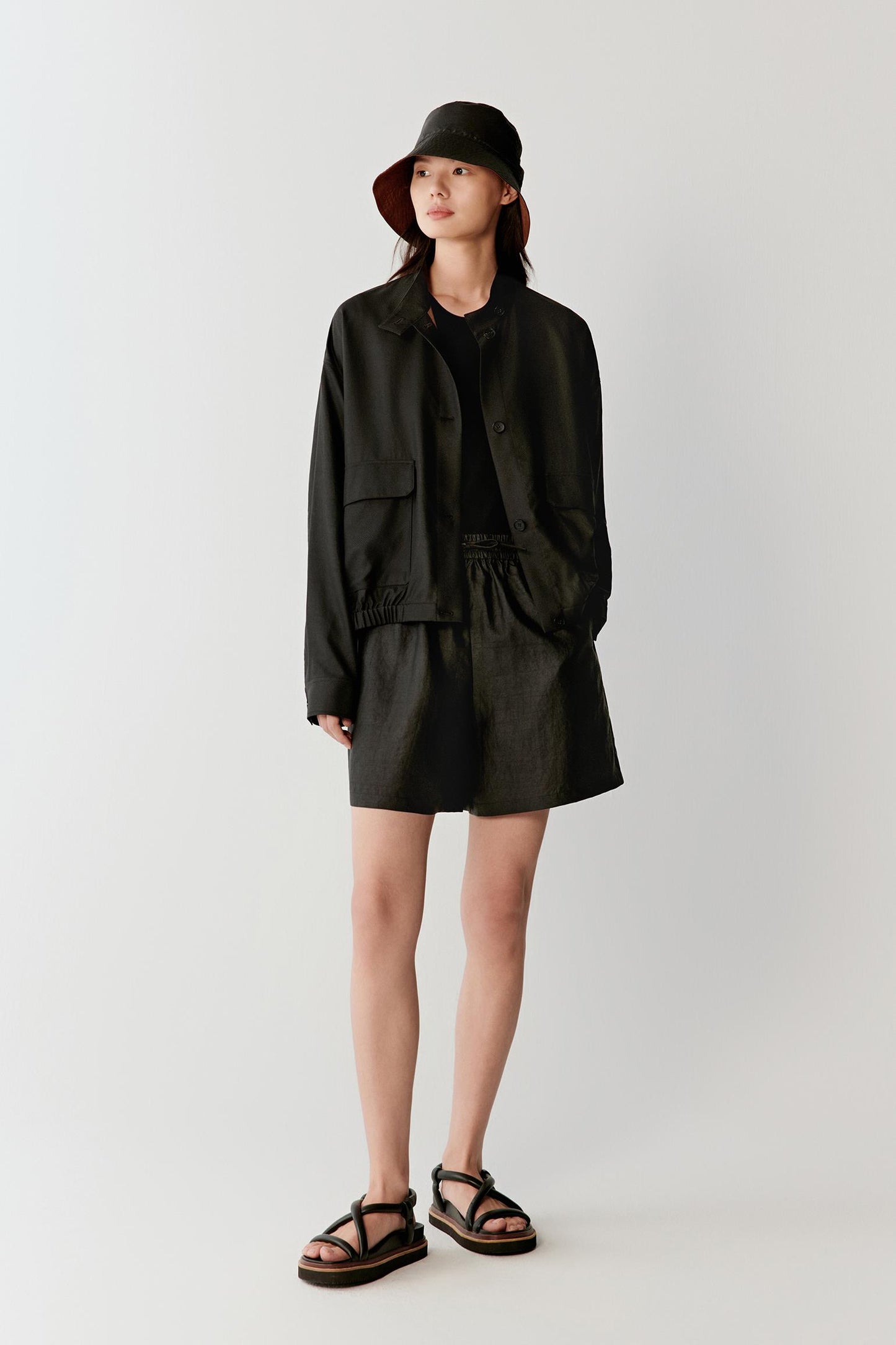 Full-length front view of a woman wearing deep black shorts in Gambiered Canton Gauze, highlighting the fluid drape, matte surface and subtle tonal depth developed through the traditional dyeing process