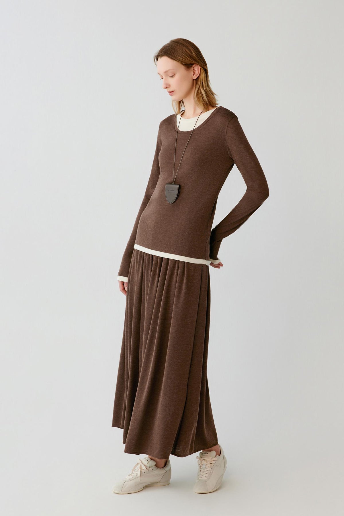 A woman modeling a brown knit long-sleeve top and matching pleated maxi skirt set with a contrasting cream hem, paired with cream sneakers and a long necklace, against a light gray studio background.