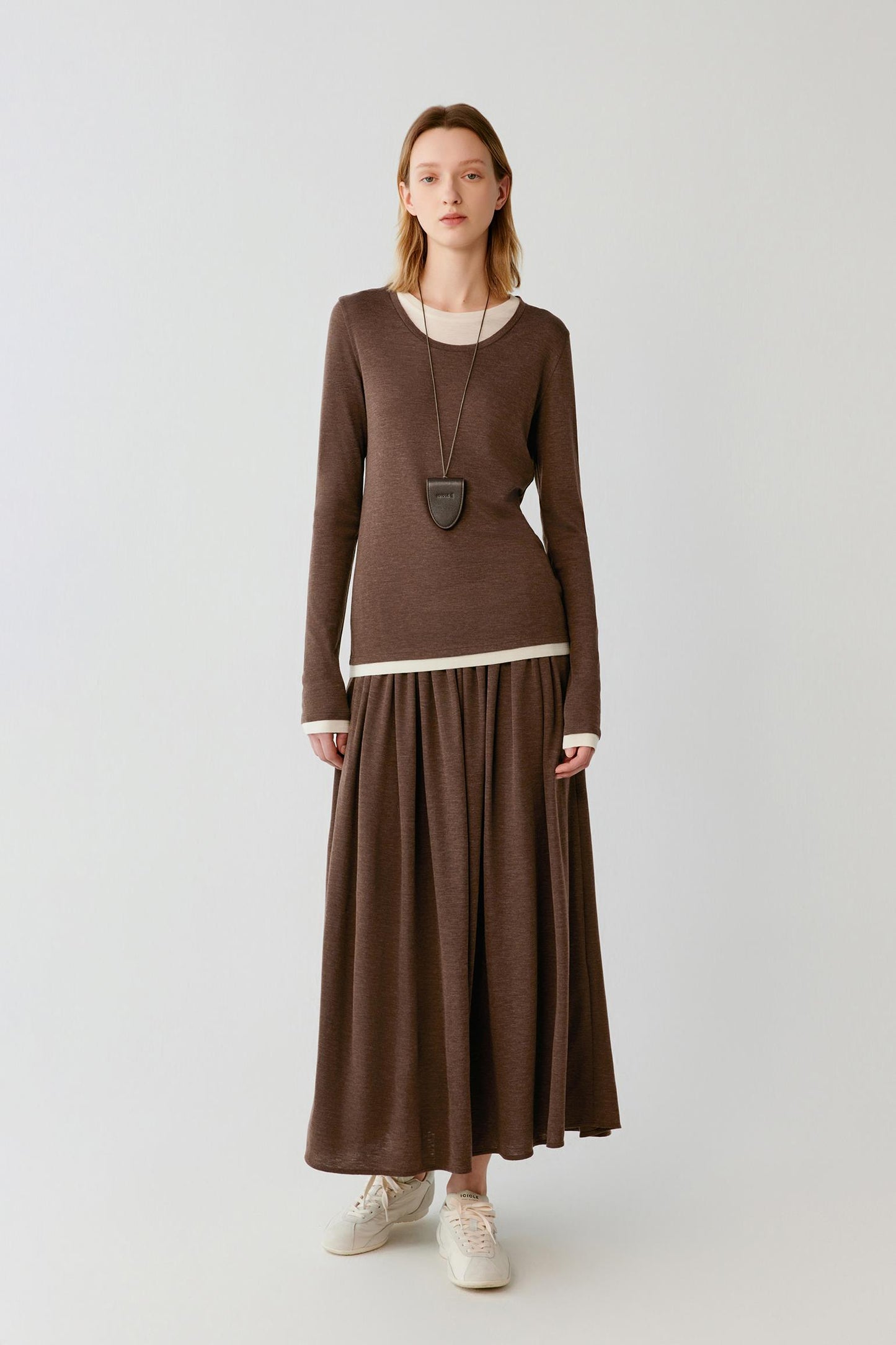 A woman stands against a light gray studio background wearing a brown long-sleeve knit top and matching pleated maxi skirt with a thin white contrast band at the waist, plus white sneakers and a long dark pendant necklace.
