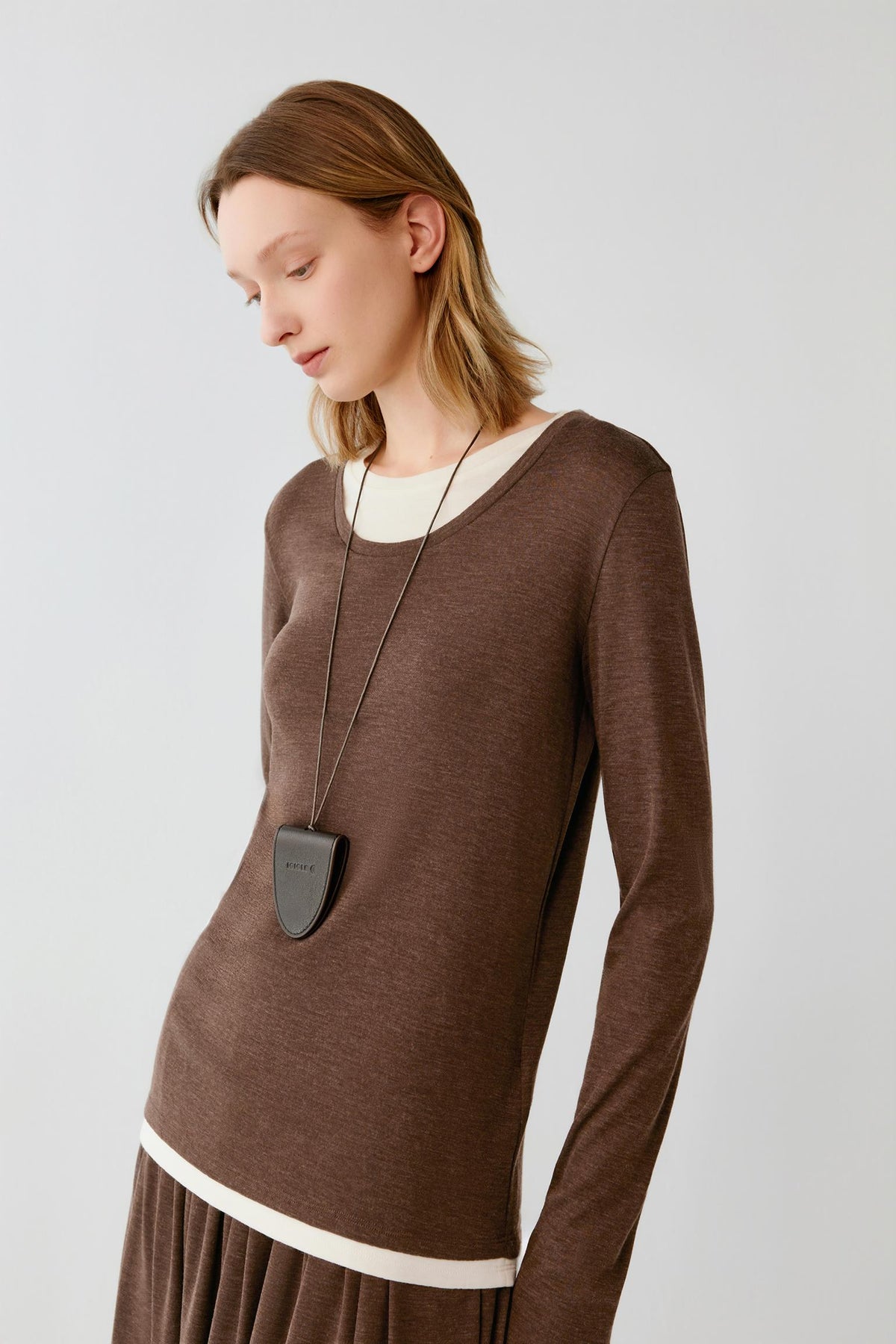 A woman wearing a brown long-sleeve knit top with contrasting cream ribbed trim at the crew neckline and hem, styled with a dark gray shield-shaped pendant necklace, against a light gray studio background.