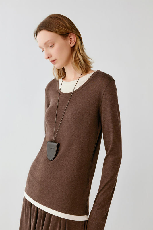 A woman wearing a brown long-sleeve knit top with contrasting cream ribbed trim at the crew neckline and hem, styled with a dark gray shield-shaped pendant necklace, against a light gray studio background.