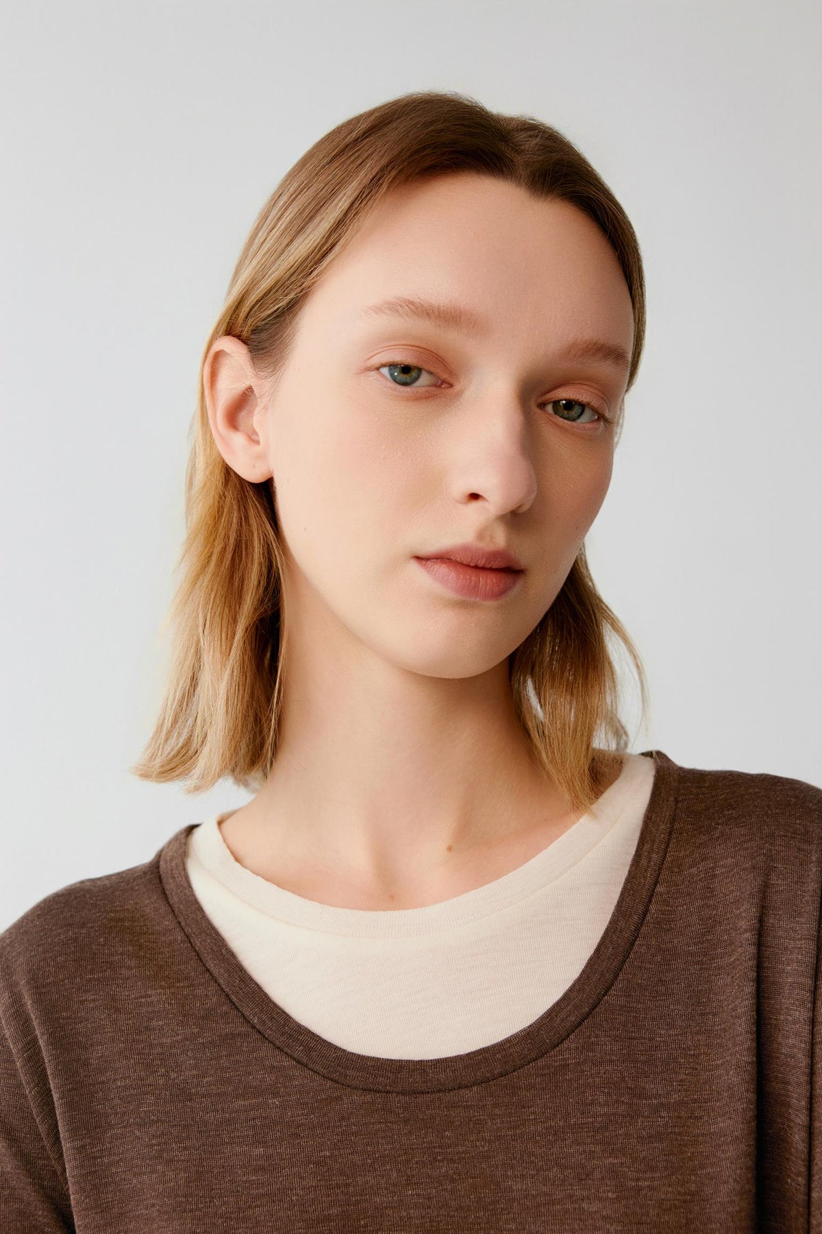A close-up portrait of a woman with shoulder-length blonde hair wearing a brown crewneck sweater layered over a cream-colored t-shirt, set against a plain light gray background.