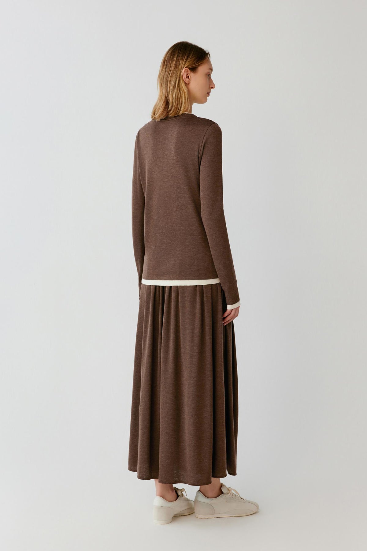 A woman shown from the back wearing a brown knit long-sleeve top and matching pleated maxi skirt with a thin cream contrast hem, paired with off-white sneakers, against a plain light gray studio background.
