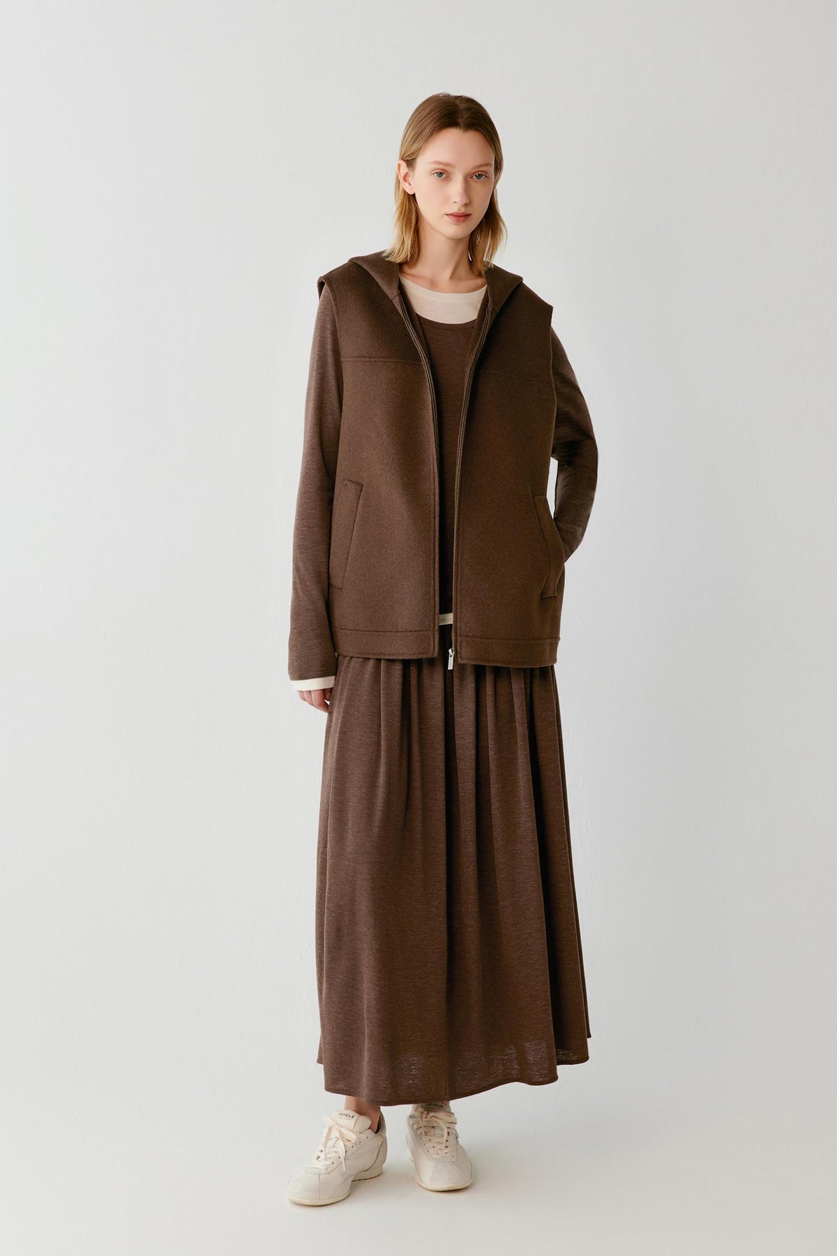 Woman wearing a brown hooded zip-up vest with a matching maxi skirt and white trainers.