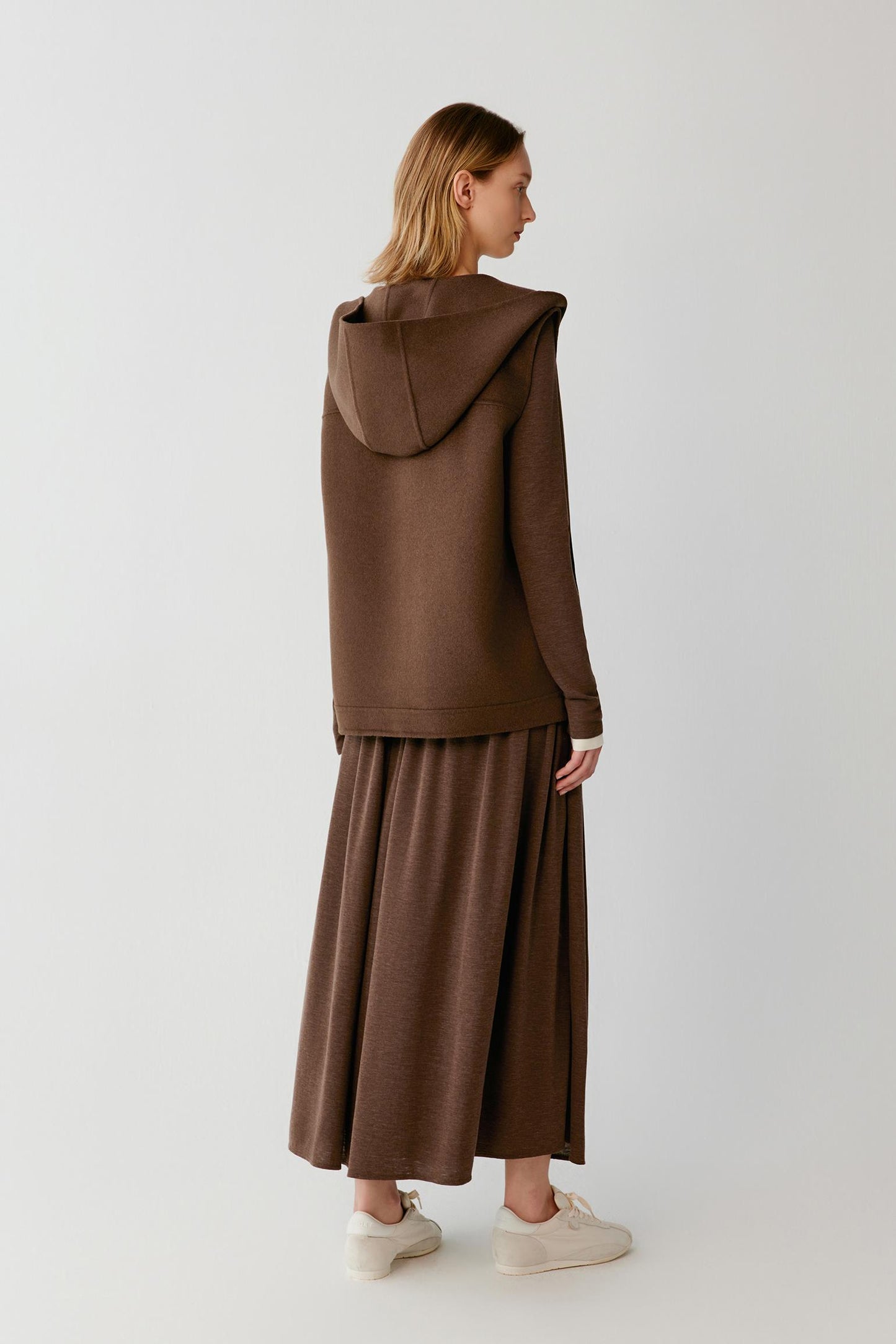 Back view of a woman wearing a brown hooded vest with a matching maxi skirt and white trainers.