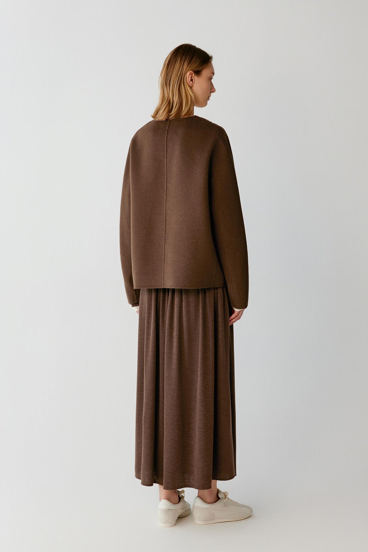 Back view of a woman wearing a brown jacket with a matching maxi skirt and white trainers.