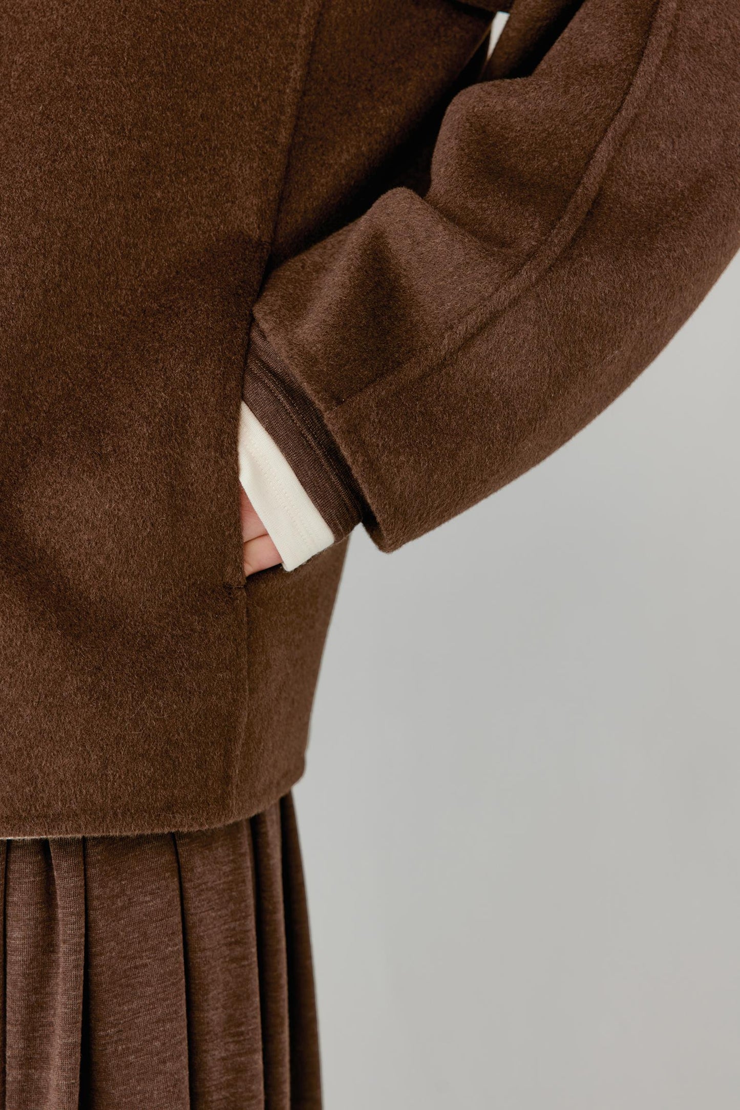 Close-up of a brown wool-like coat with a side pocket, showing a hand tucked inside, paired with a matching brown pleated skirt against a light gray background.