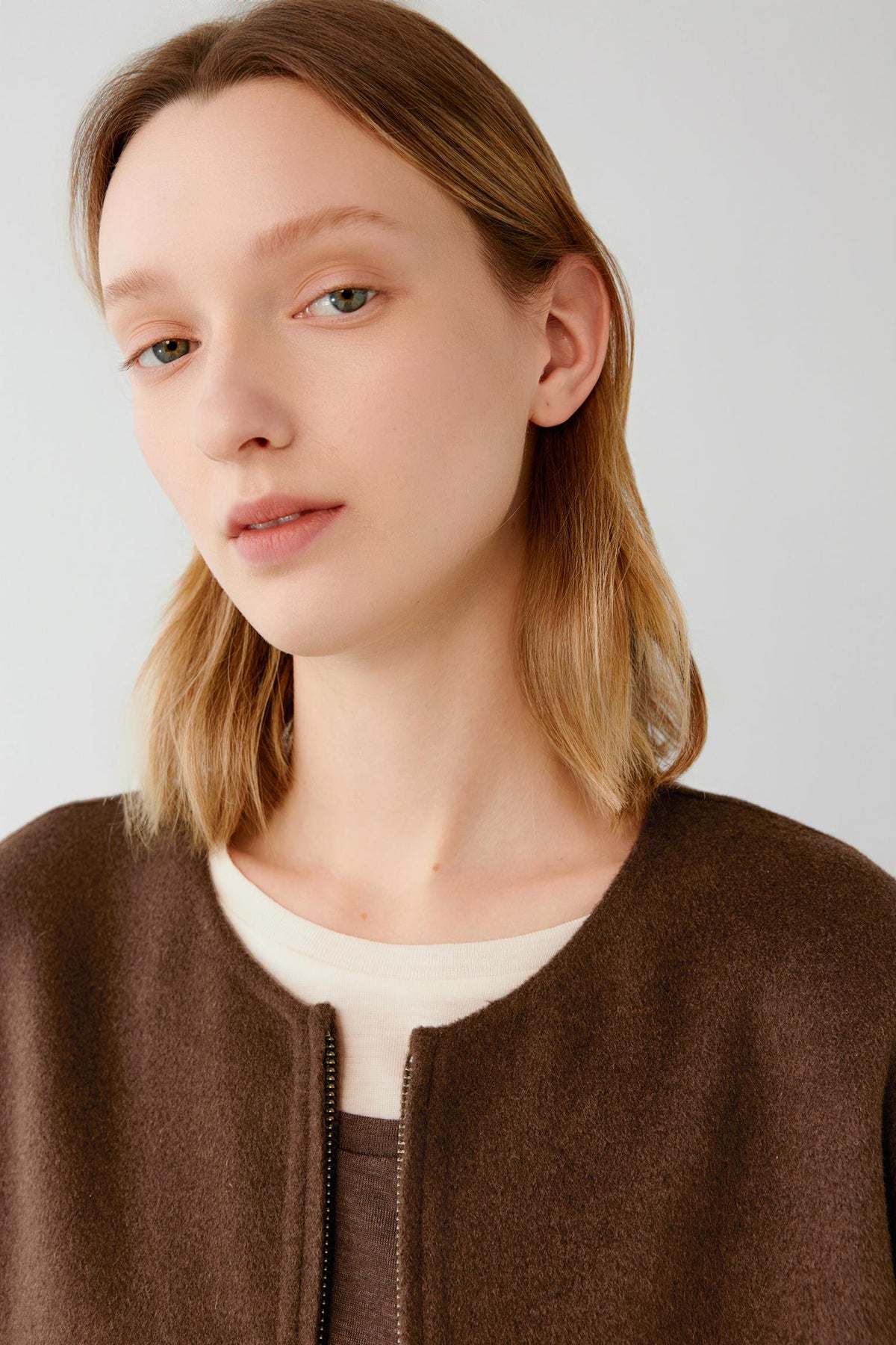 A close-up portrait of a woman with light brown hair wearing a brown zip-up jacket over a cream-colored top, set against a plain light gray background.