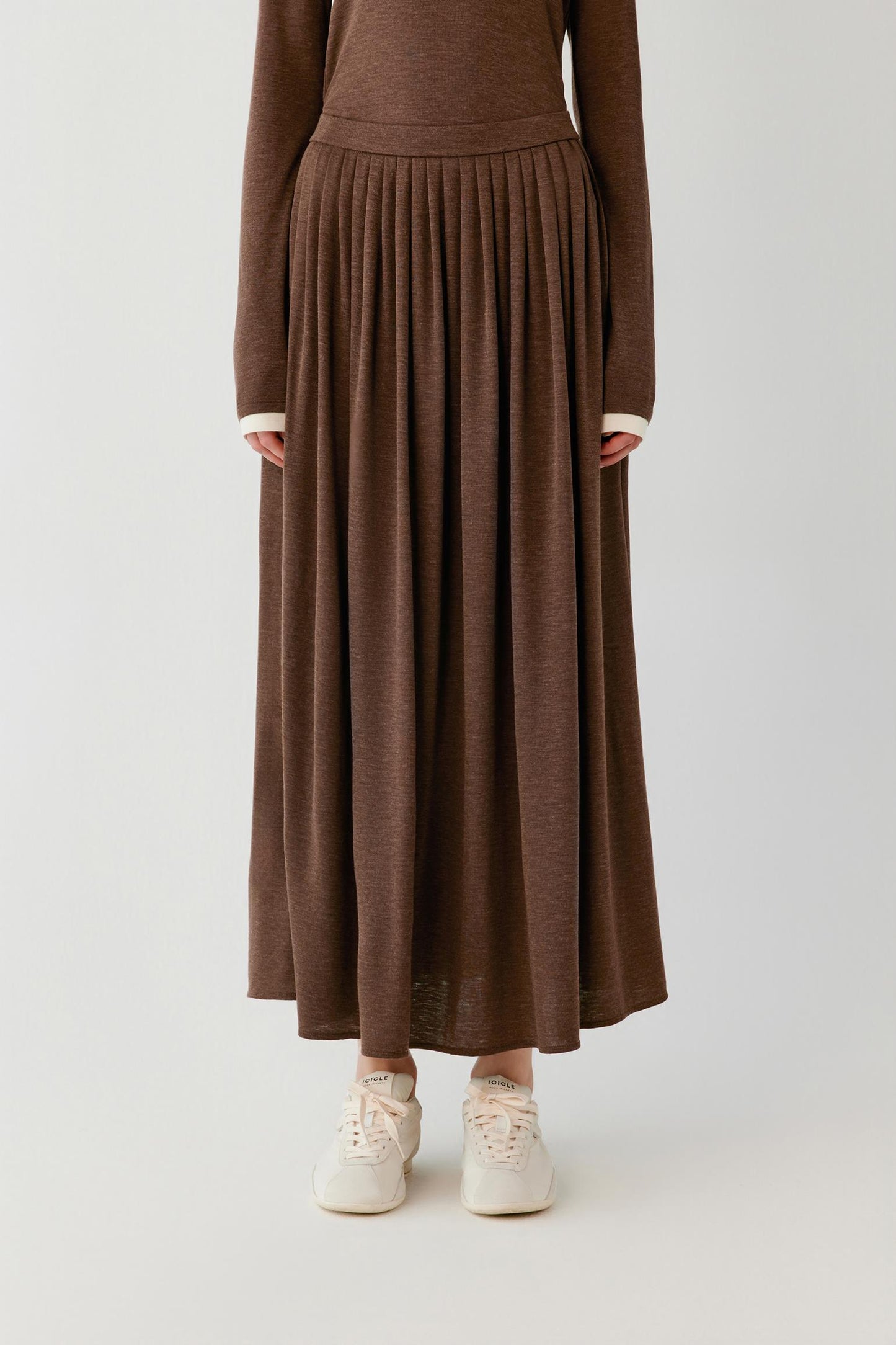 A person wearing a long-sleeve brown knit maxi dress with a pleated skirt and white sneakers, photographed against a plain light gray studio background.